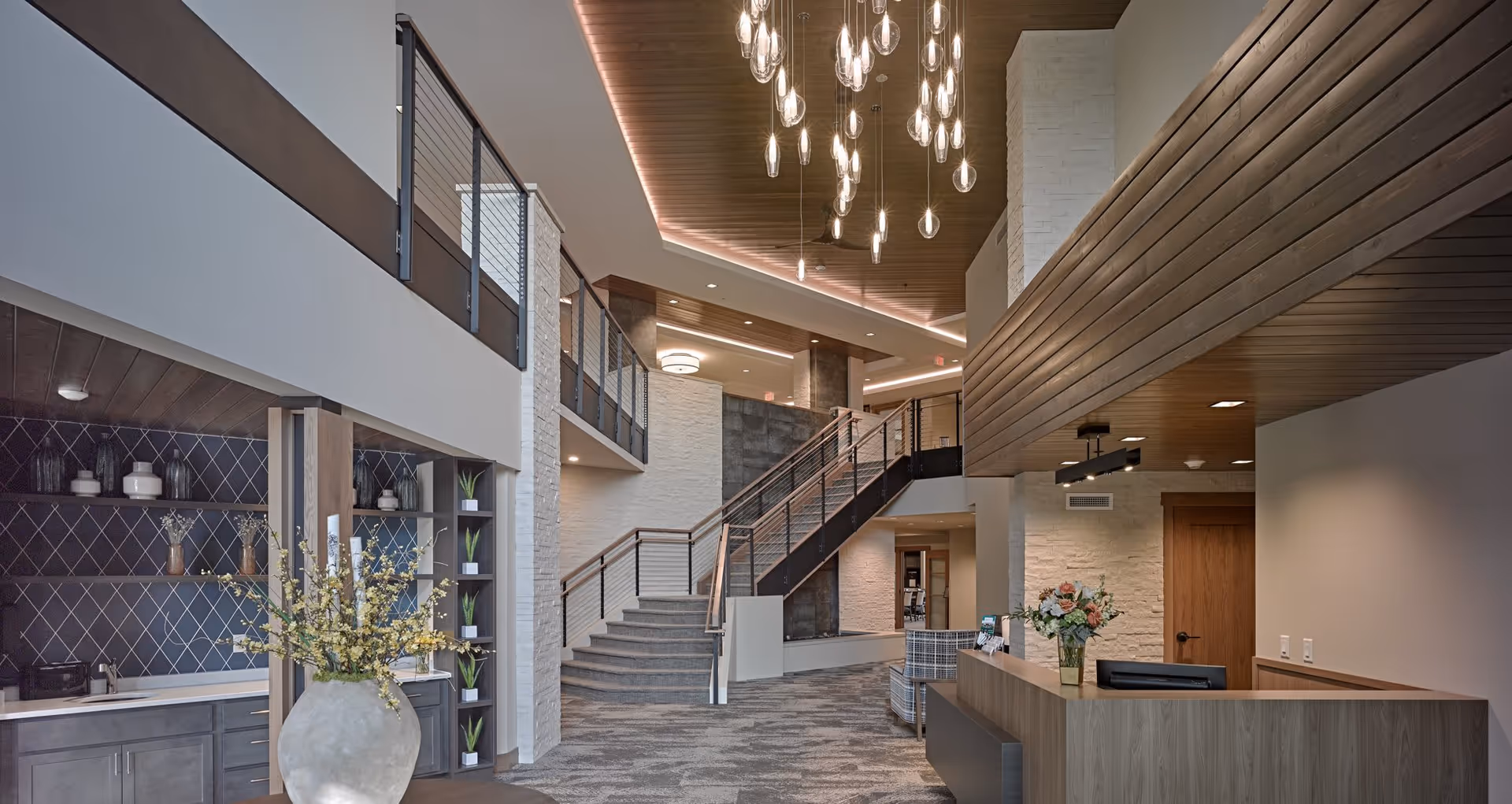 Modern senior living facility interior with a reception desk on the right, a large vase with flowers on the left, a staircase leading to an upper floor, and contemporary hanging light fixtures from a wooden ceiling.