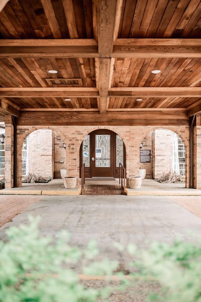 Covered entrance area with wooden ceiling beams and brick arches leading to a glass door with decorative metalwork. There are two large planters on either side of the entrance and some greenery in the foreground.