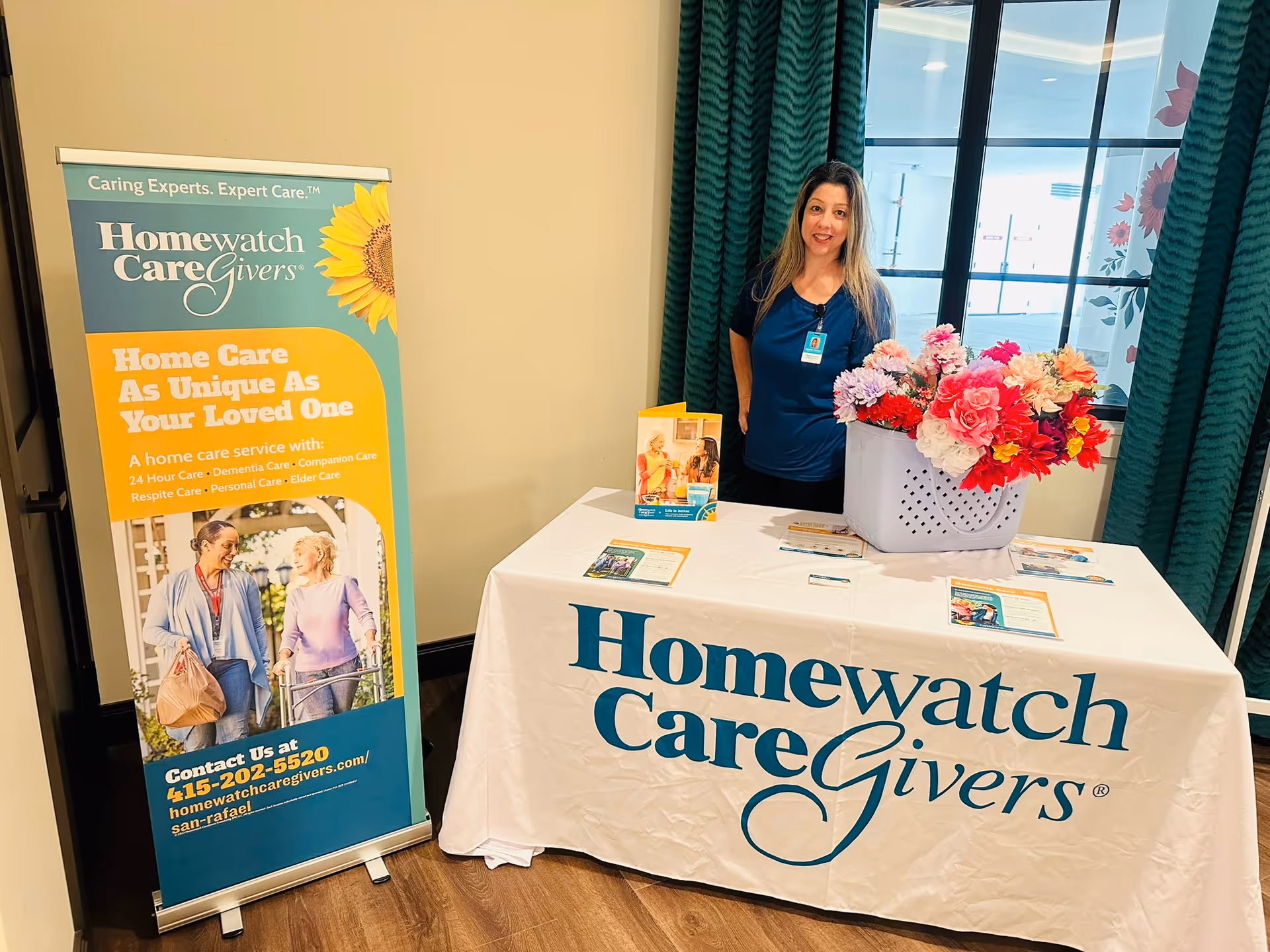 A staff member stands beside a Homewatch CareGivers promotional banner indoors.