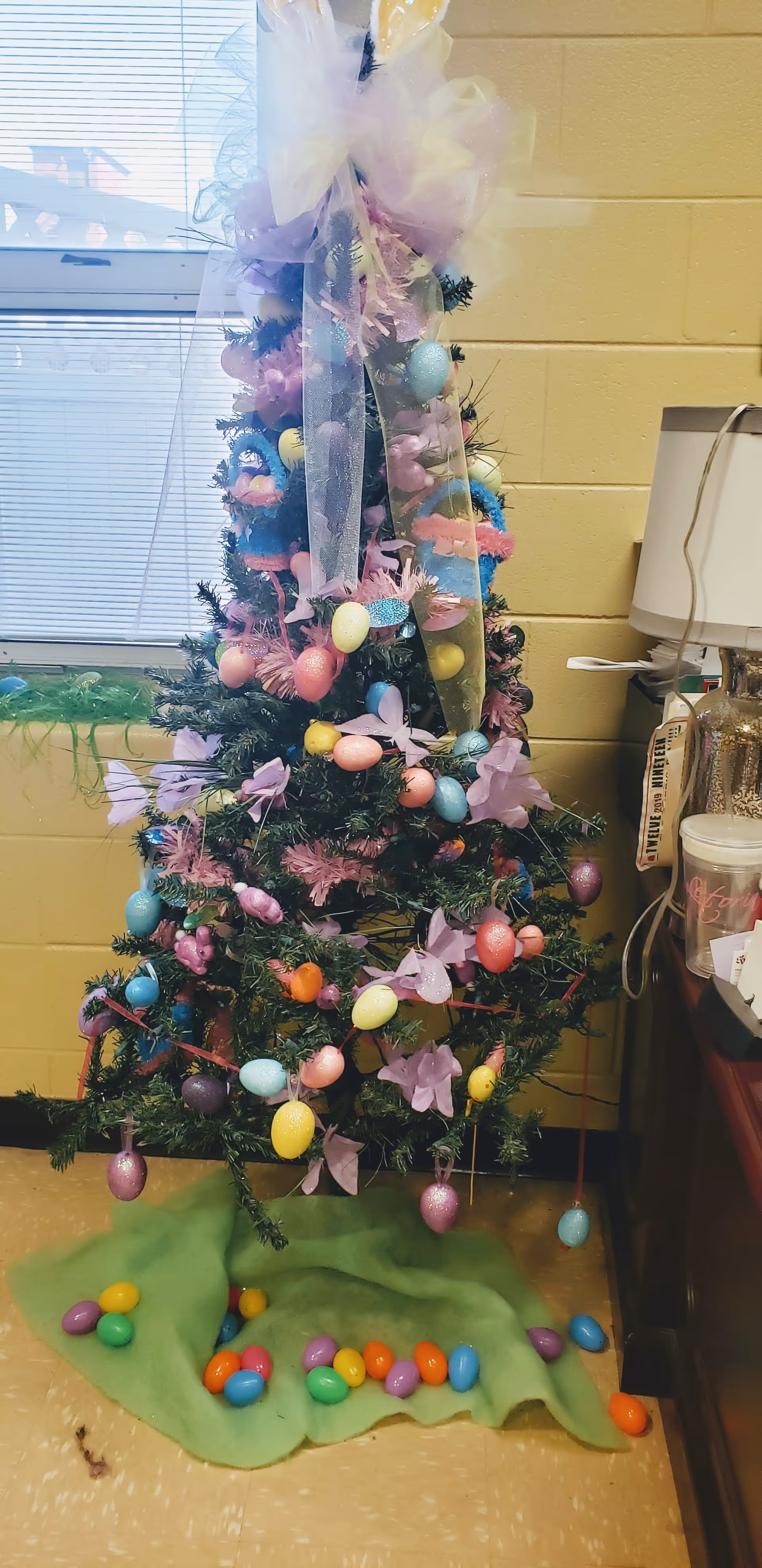 Small decorated tree with pastel Easter egg ornaments, ribbons and scattered plastic eggs on a green cloth next to a window and lamp.