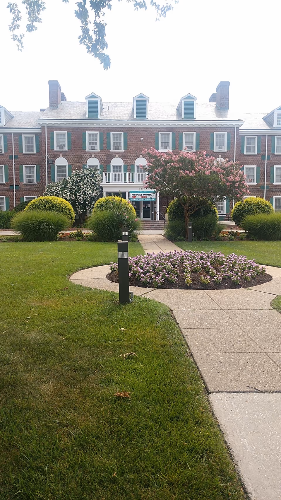 Brick senior living building with green shutters, manicured lawn, flowering trees, and walkway to the entrance.