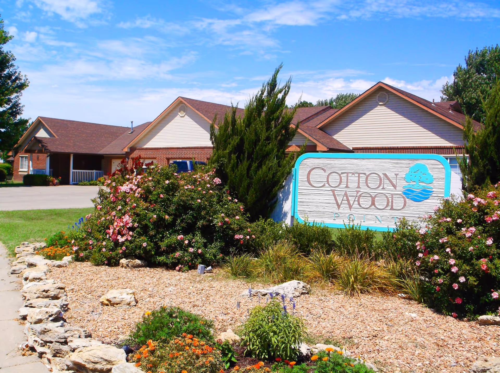 A landscaped entrance with a 'Cotton Wood' sign in front of a single-story brick senior living building under a blue sky.