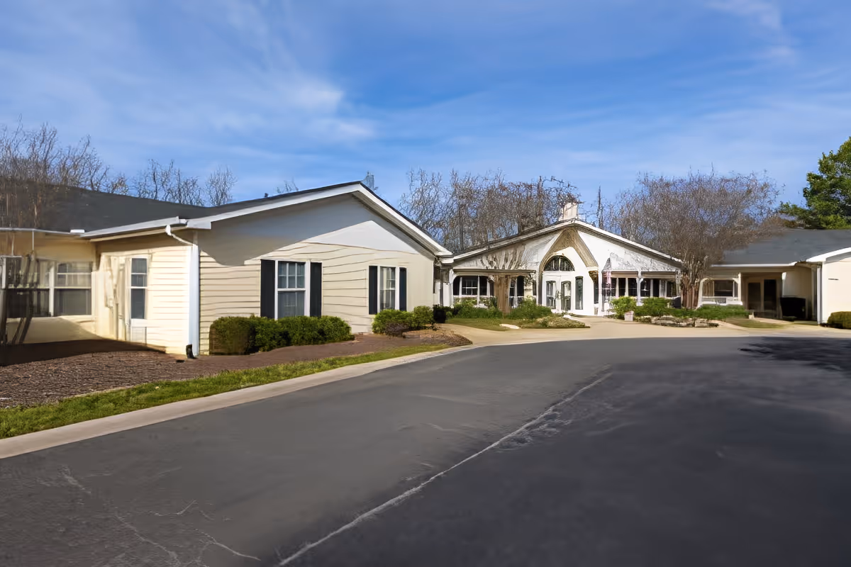 Front exterior of a single-story senior living facility with landscaped grounds and a circular driveway under a blue sky.