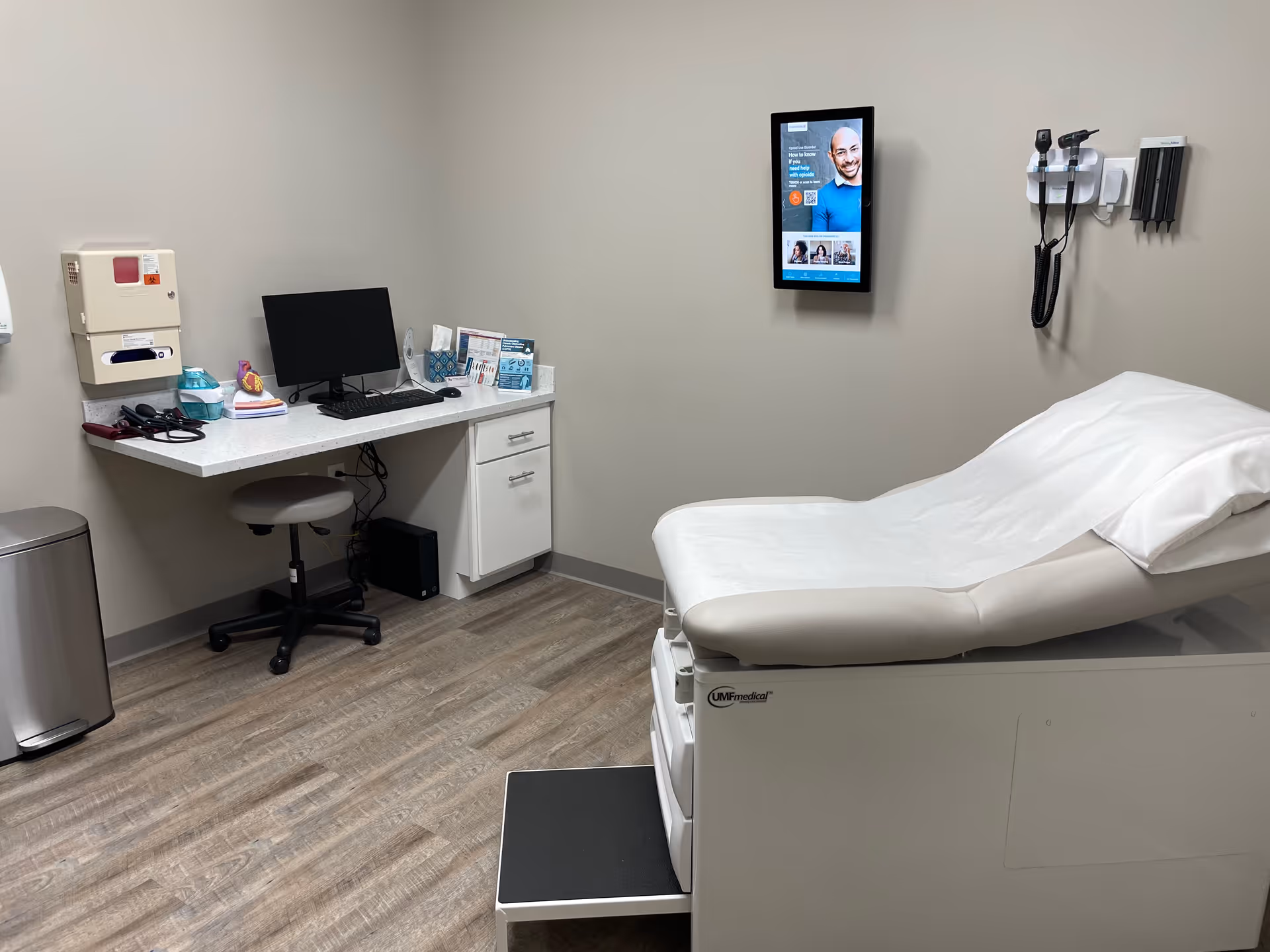 Clinical examination room with a medical bed, computer desk, and wall-mounted equipment.