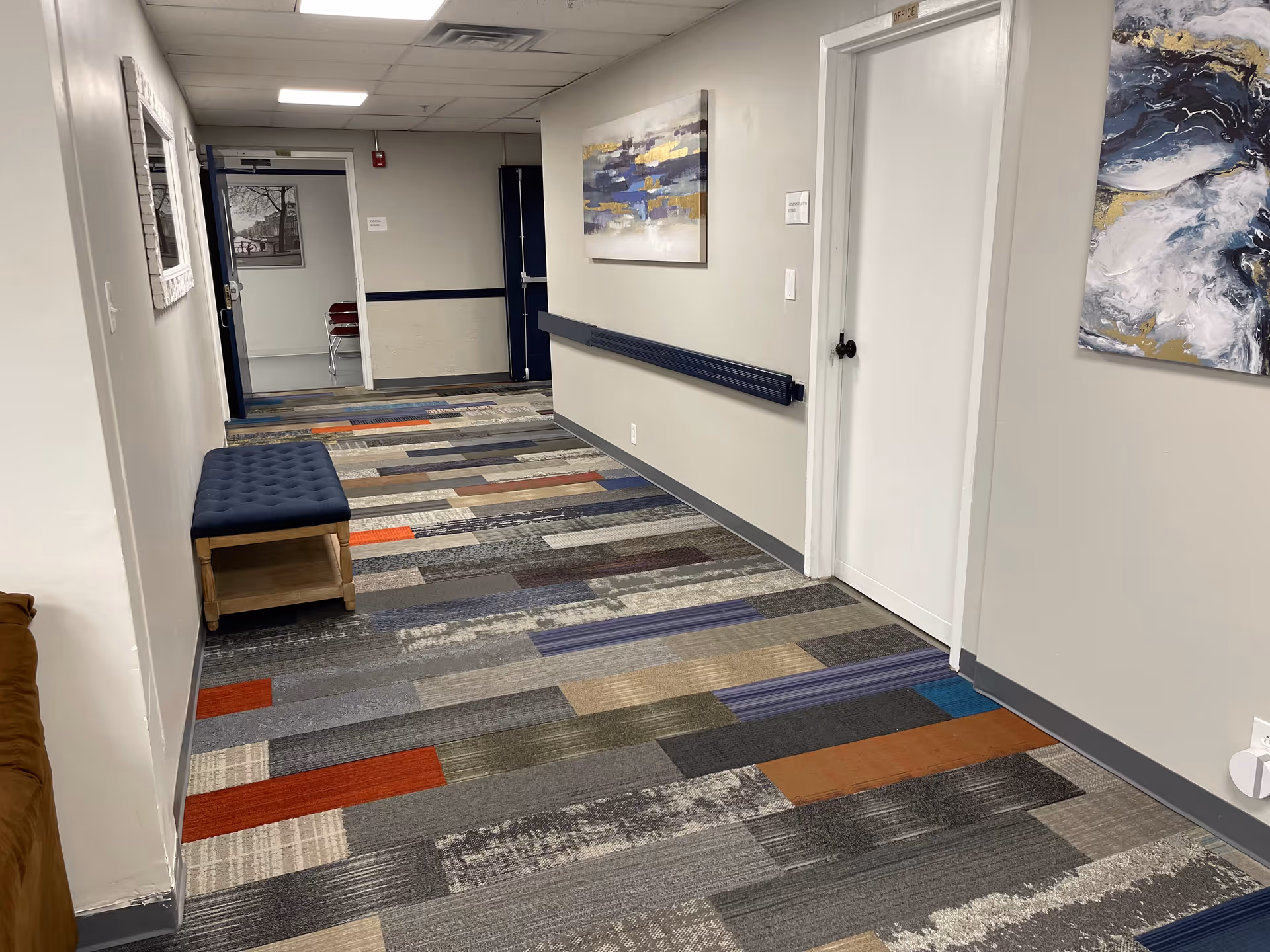 Well-lit interior hallway in a senior living facility with colorful carpet, bench, and artwork.