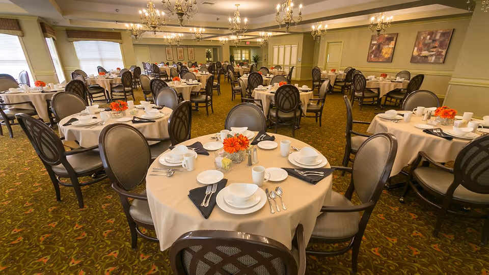 A spacious dining room in a senior living facility with multiple round tables covered with beige tablecloths, each set with white plates, bowls, cups, silverware, black napkins, and small orange floral centerpieces. The room has green patterned carpet, green walls with framed artwork, and large windows letting in natural light.