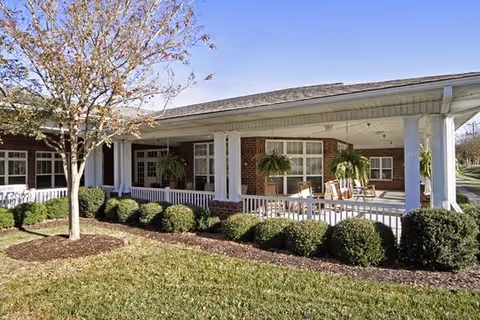Exterior view of a senior living facility with a covered porch featuring rocking chairs and hanging plants. The building has brick walls and white columns, with neatly trimmed bushes and a tree in the foreground under a clear blue sky.