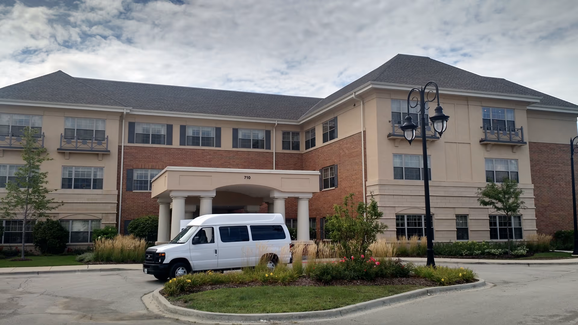Three-story beige brick senior living building with an entrance and a white van out front.