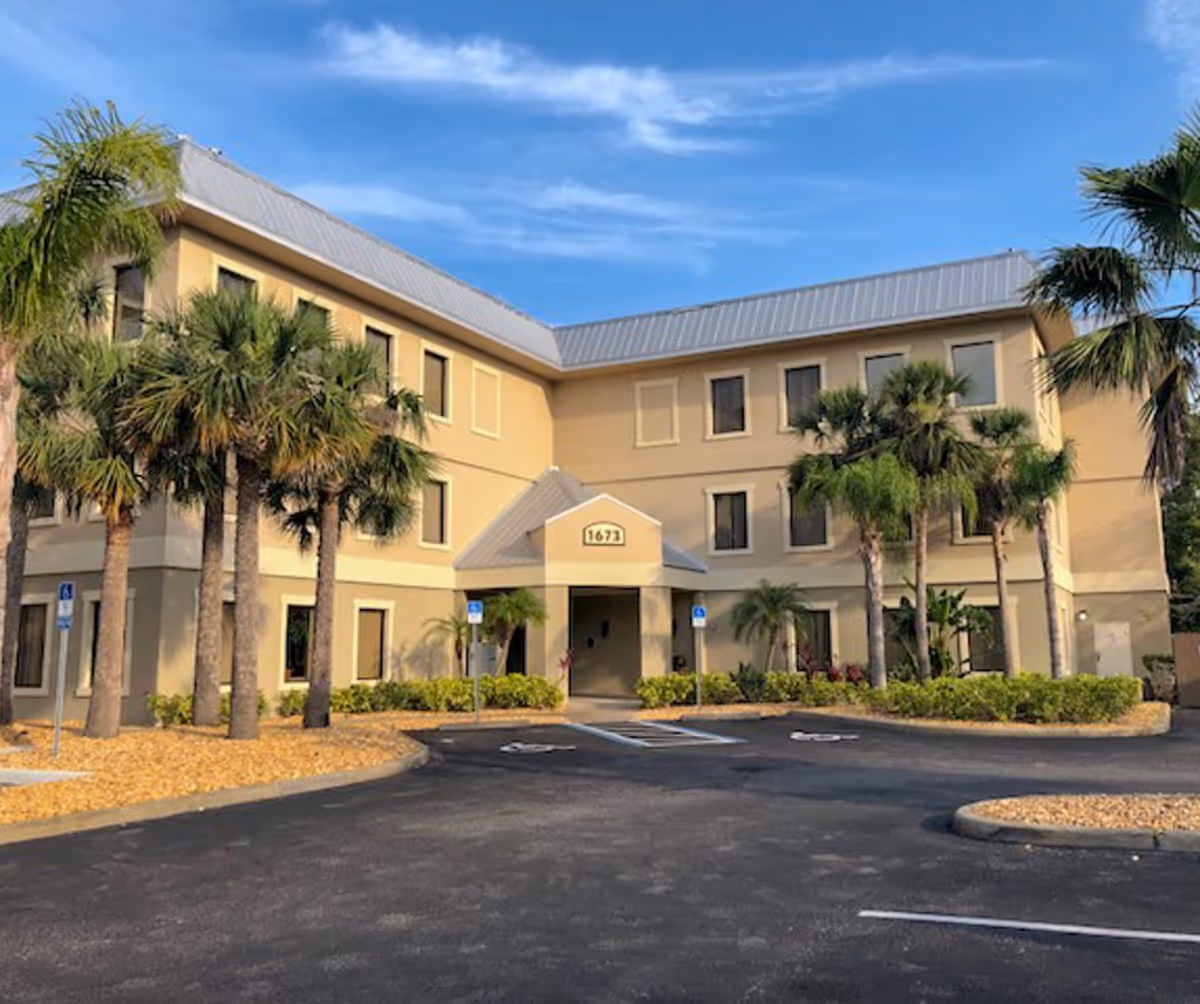 Two-story beige senior living building with palm trees and a paved entrance under a blue sky.