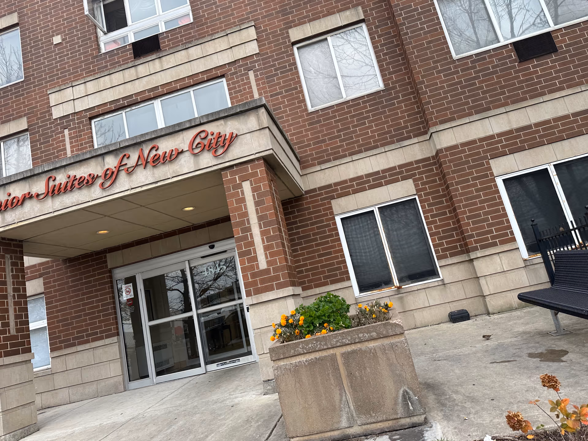 Entrance of a brick senior living building with a canopy sign reading 'Senior Suites of New City' above glass double doors and a planter with flowers.