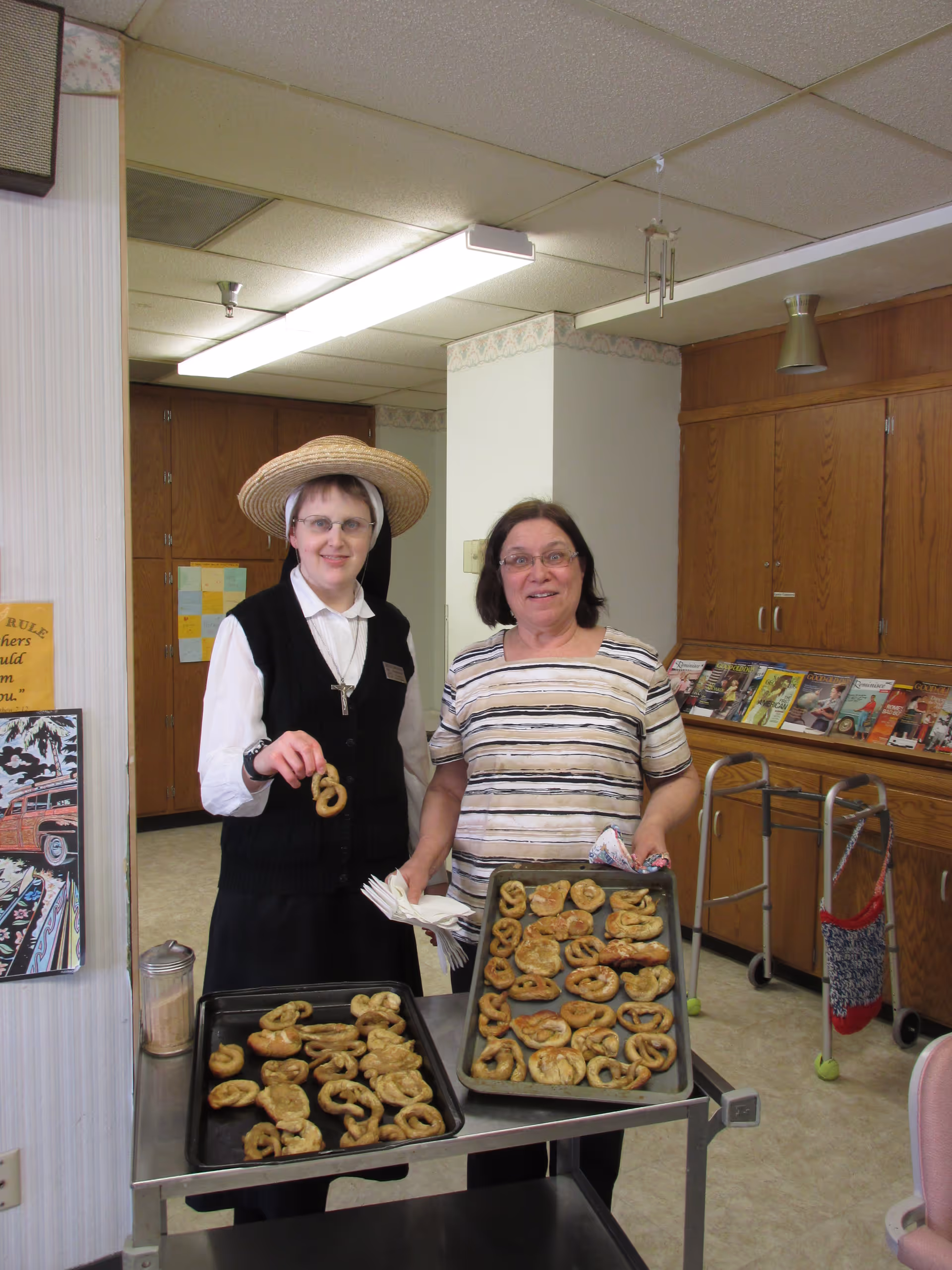 Two women standing indoors in a room with wooden cabinets and a walker in the background. One woman is wearing a black vest, white shirt, and a straw hat, holding a pretzel. The other woman is wearing glasses and a striped shirt, holding a baking tray filled with pretzels. Another tray with pretzels is on a metal cart in front of them.