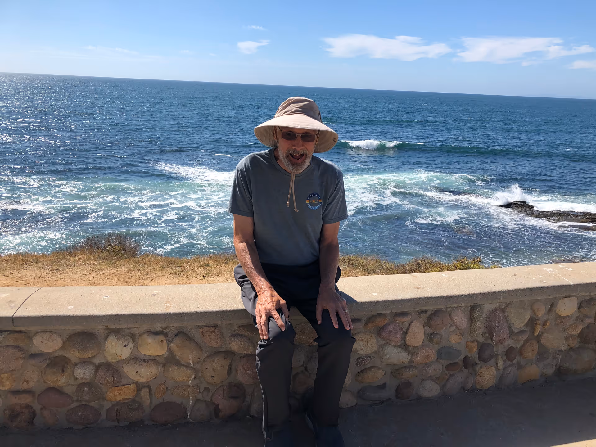 An elderly man wearing a wide-brimmed hat, sunglasses, a gray t-shirt, and dark pants is sitting on a stone wall with the ocean and waves visible in the background under a clear blue sky.