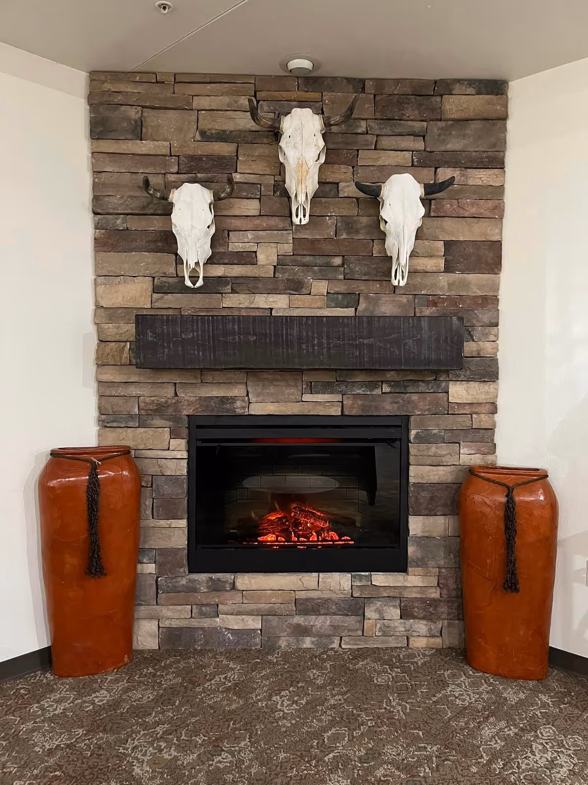 A stone fireplace with a dark wooden mantel, featuring three white animal skulls with horns mounted on the stone wall above it. On either side of the fireplace, there are tall, brown decorative vases with black tassels. The floor is carpeted with a patterned design.