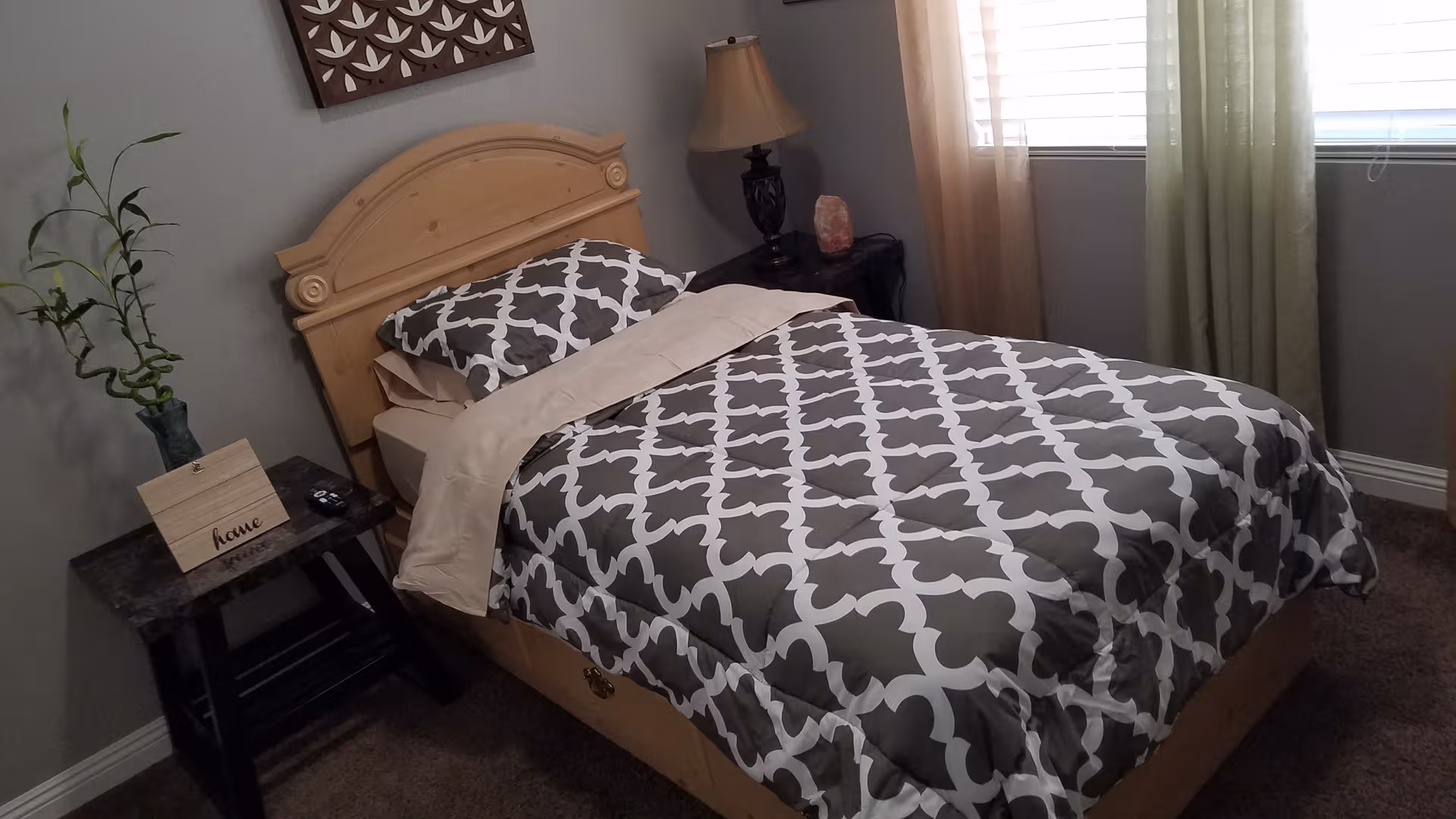 Light wood bed with gray geometric comforter, bedside tables, lamp, and plant in a bedroom.