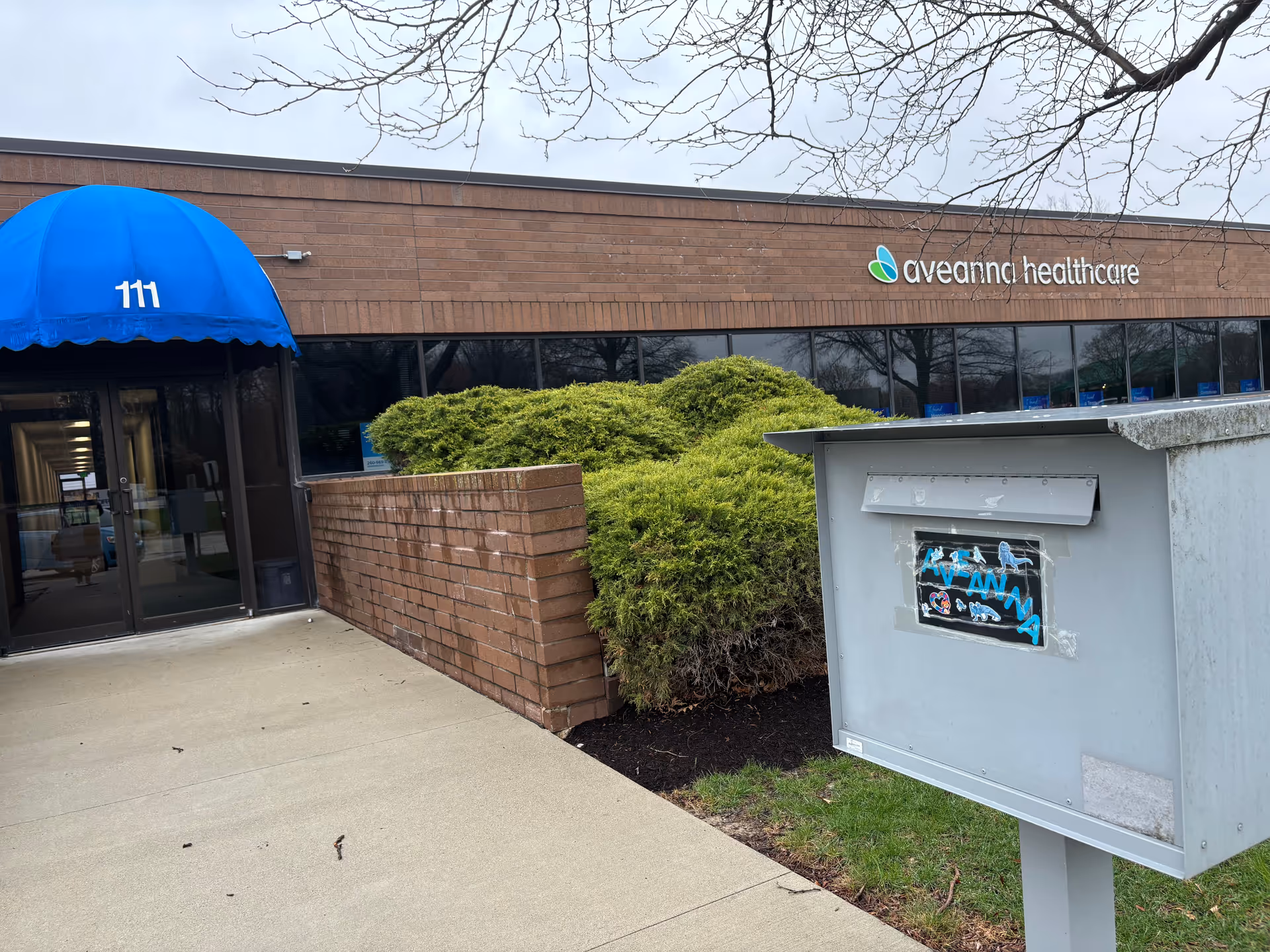 Exterior of Aveanna Healthcare facility with blue canopy, brick wall, and mailbox.