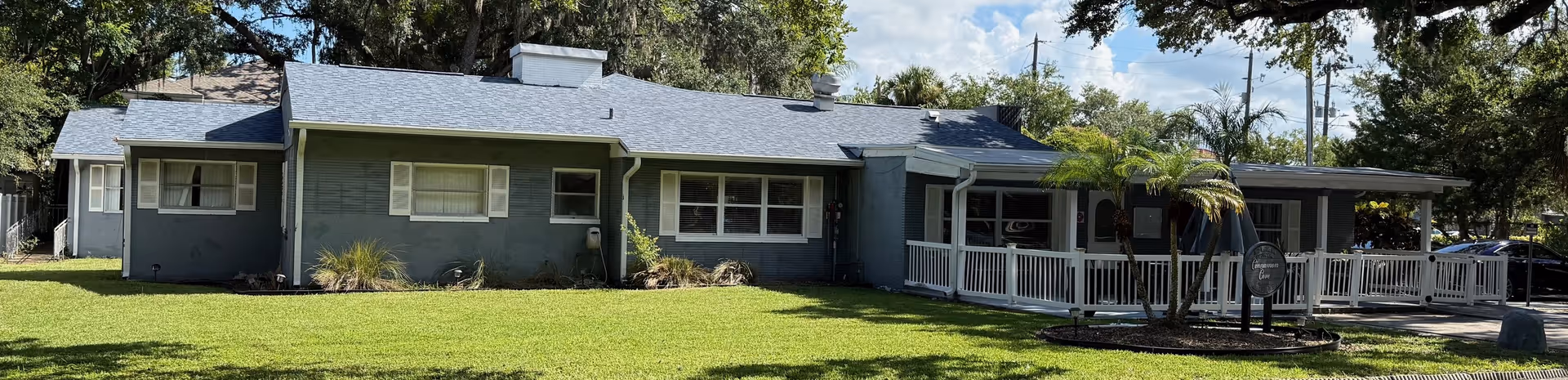 Single-story gray building with white trim and a covered porch area surrounded by a green lawn and trees under a partly cloudy sky.