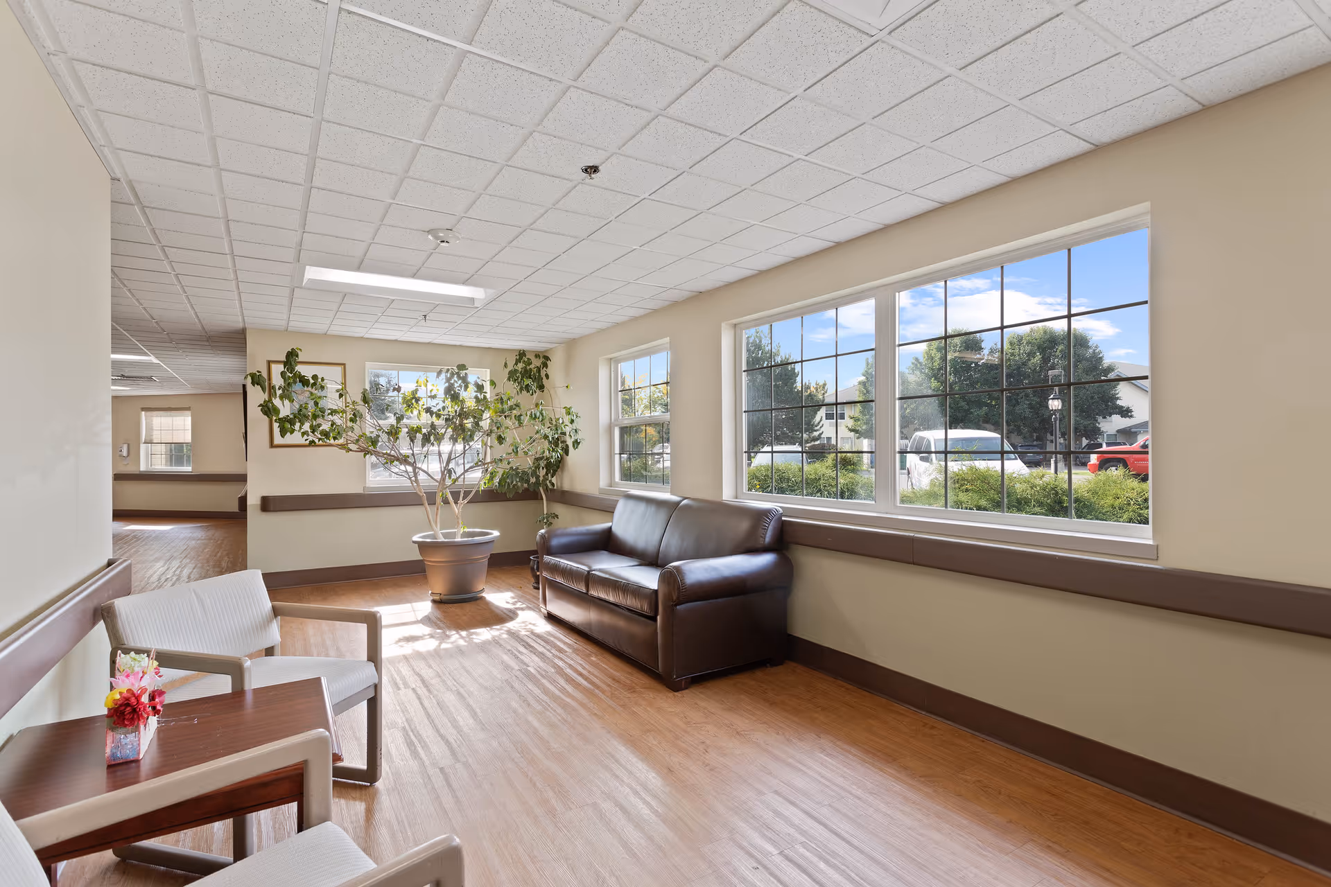 Bright seating area with a leather sofa, chairs, a side table with flowers, potted plant, and large windows looking outside.