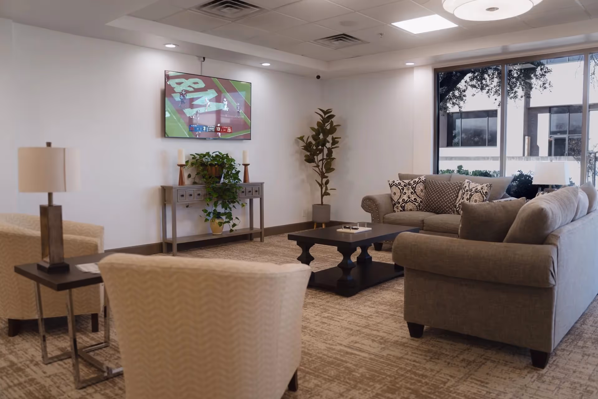 Modern assisted living lounge with sofas, chairs, plants, and a wall-mounted TV.