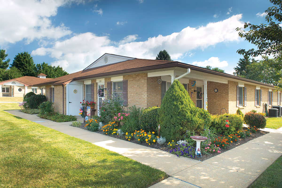 A single-story brick senior living building with colorful flower beds along a walkway.