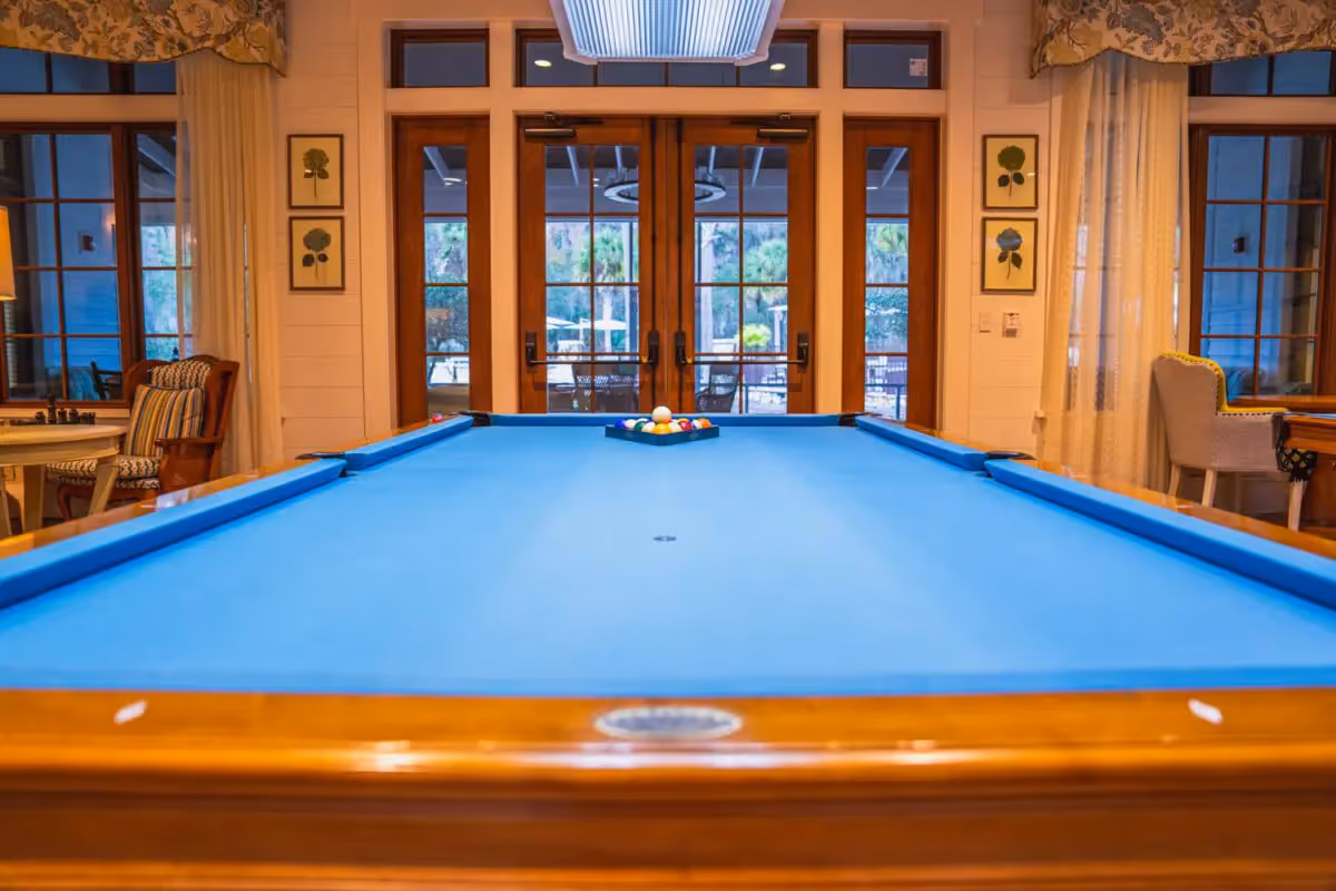An indoor recreation room with a bright blue pool table, seating along the sides, and large glass doors at the back.