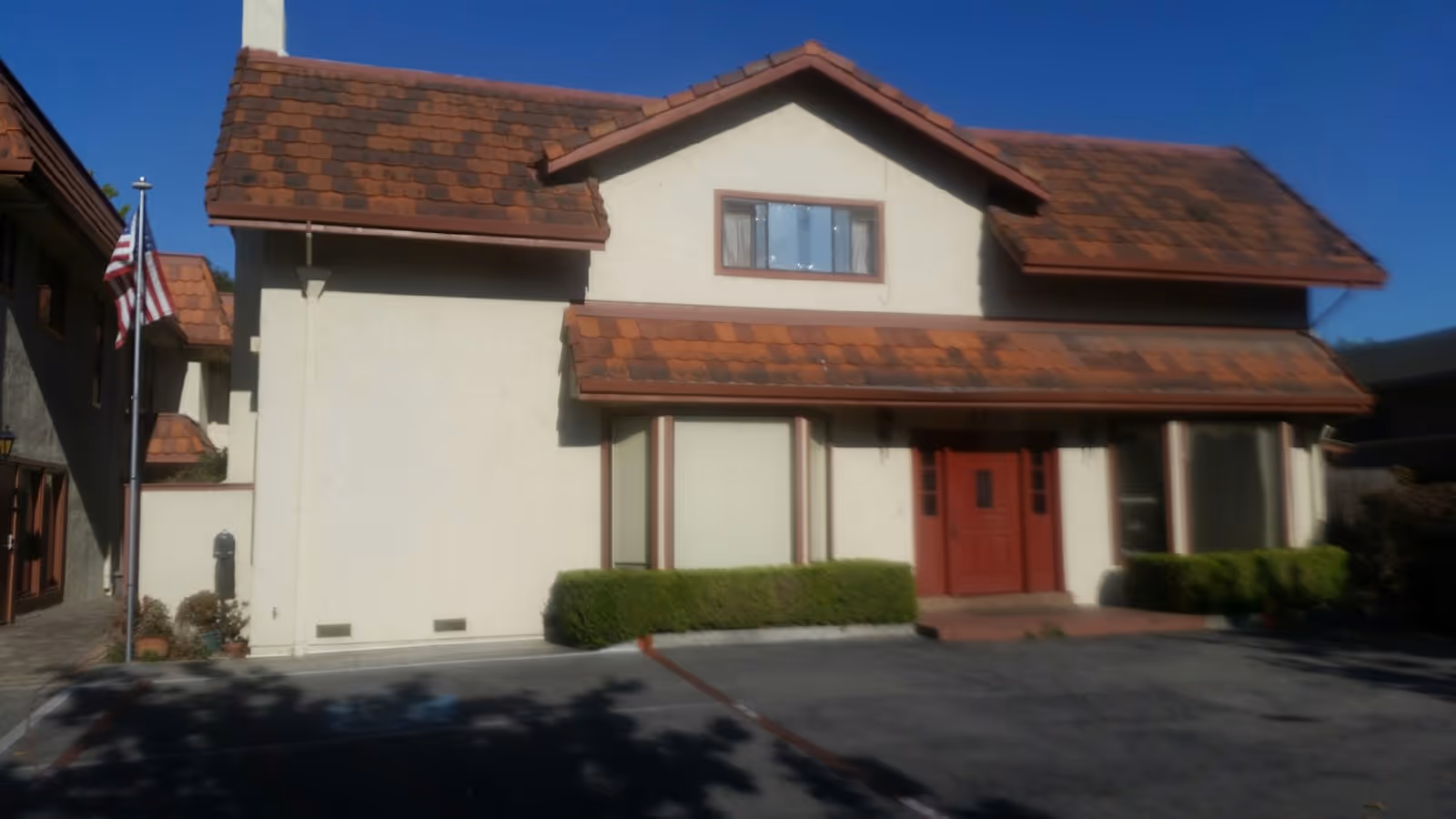 Front view of a two-story beige building with a red double door, brown tiled roof, small shrubs, a parking area, and an American flag on a pole.