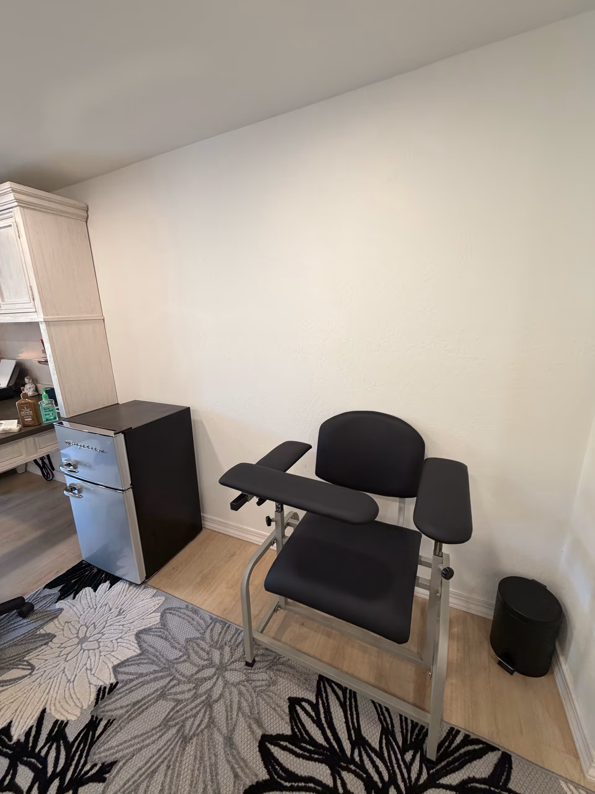 Small interior treatment room with a black reclining chair, fridge, cabinets, and rug.