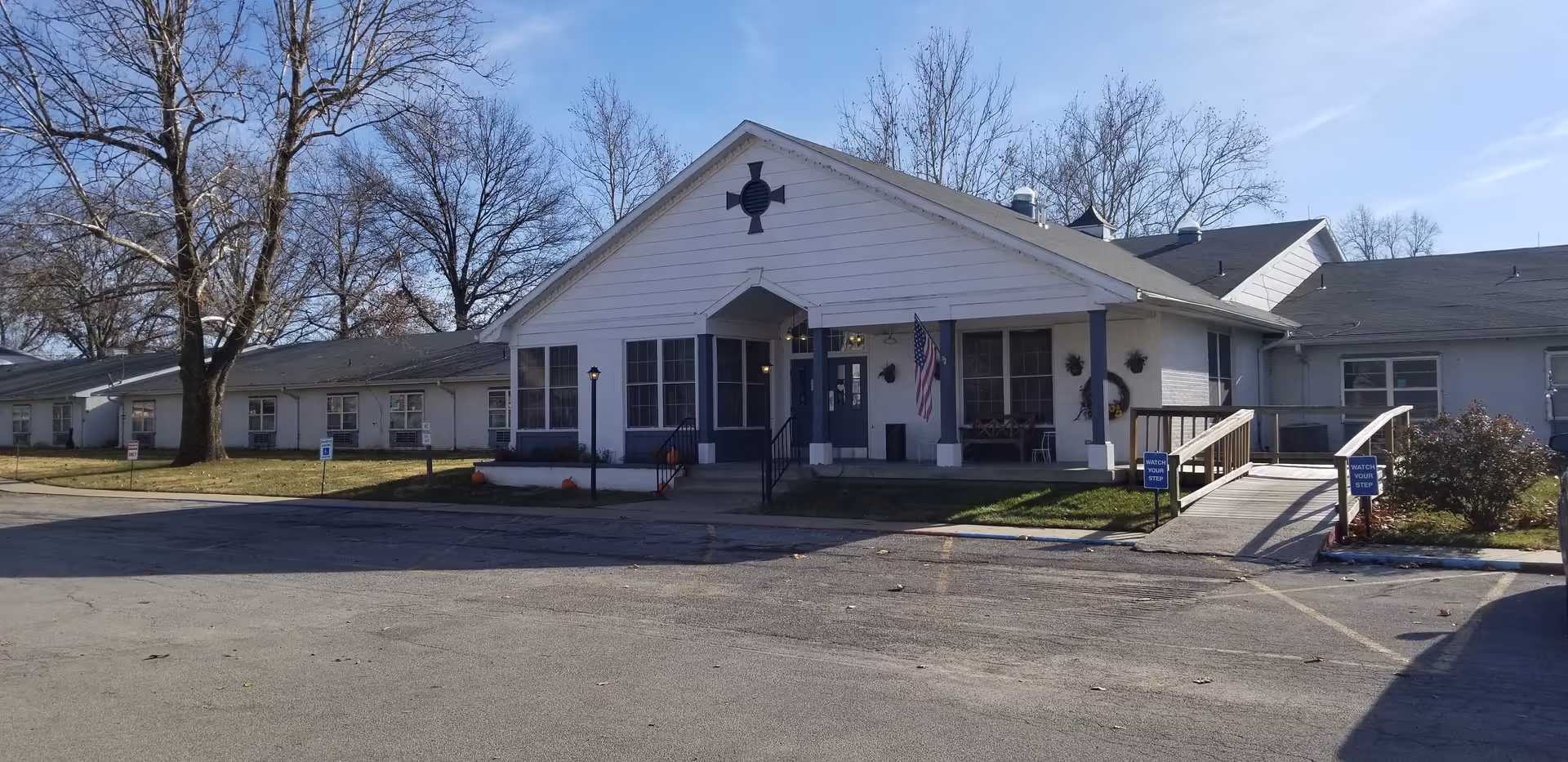 Single-story white facility with front entrance, ramp, flag, and surrounding lawn.