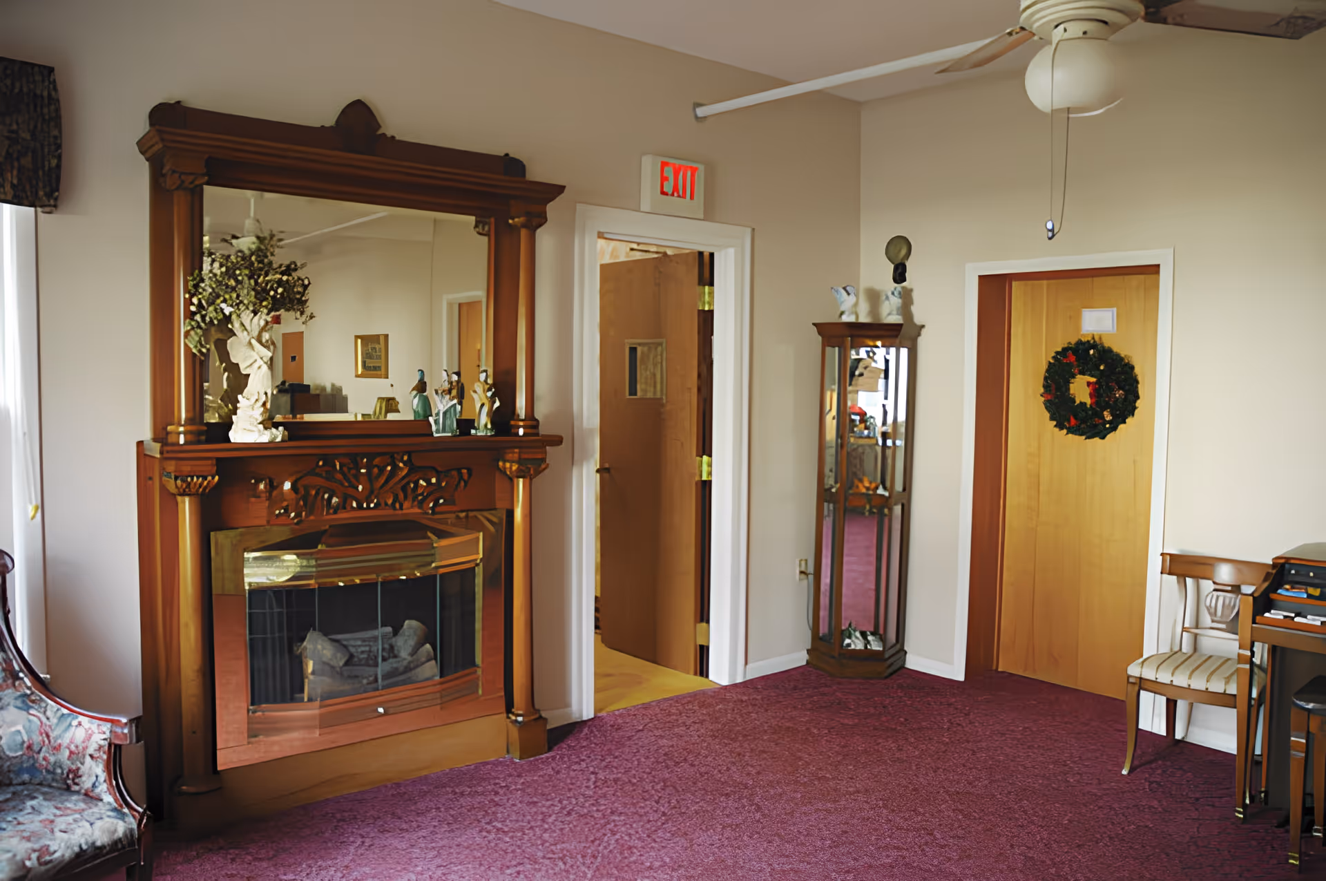 A cozy interior room with a wooden fireplace and large mirror above it, decorated with figurines and a vase of flowers. There is a floral upholstered chair on the left, a wooden display cabinet with glass shelves in the corner, and a wooden door with a wreath hanging on it. The floor is covered with a purple carpet, and a ceiling fan is visible above.