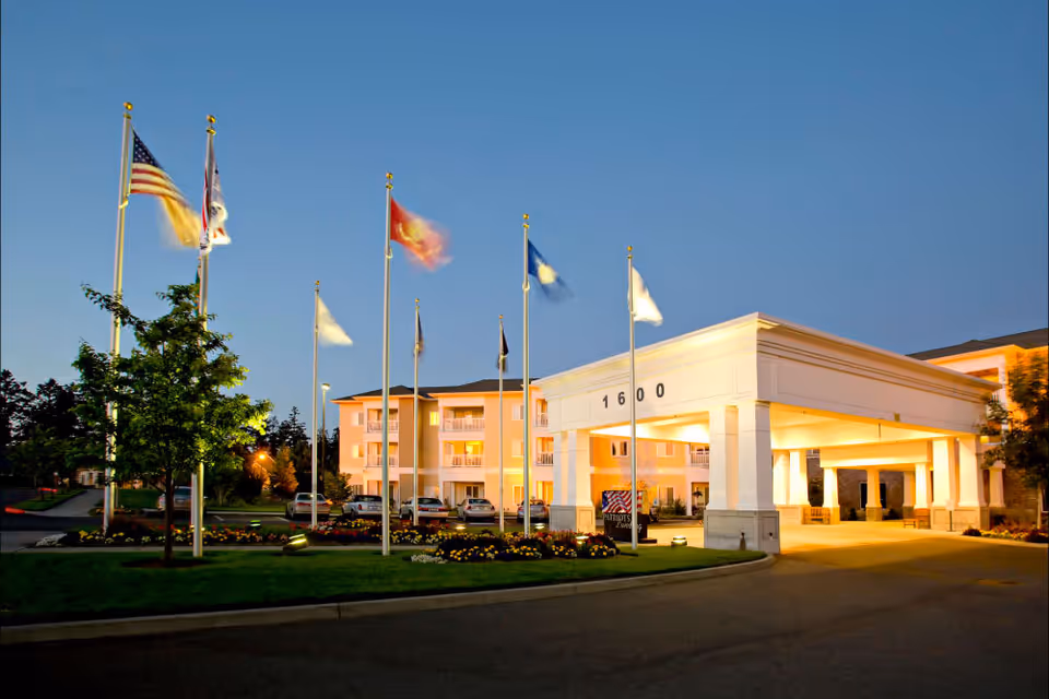 Exterior view of Patriot's Landing senior living facility at dusk, showing a large white entrance canopy with the number 1600, multiple flagpoles with flags, a landscaped area with flowers and trees, and a multi-story building with balconies in the background.