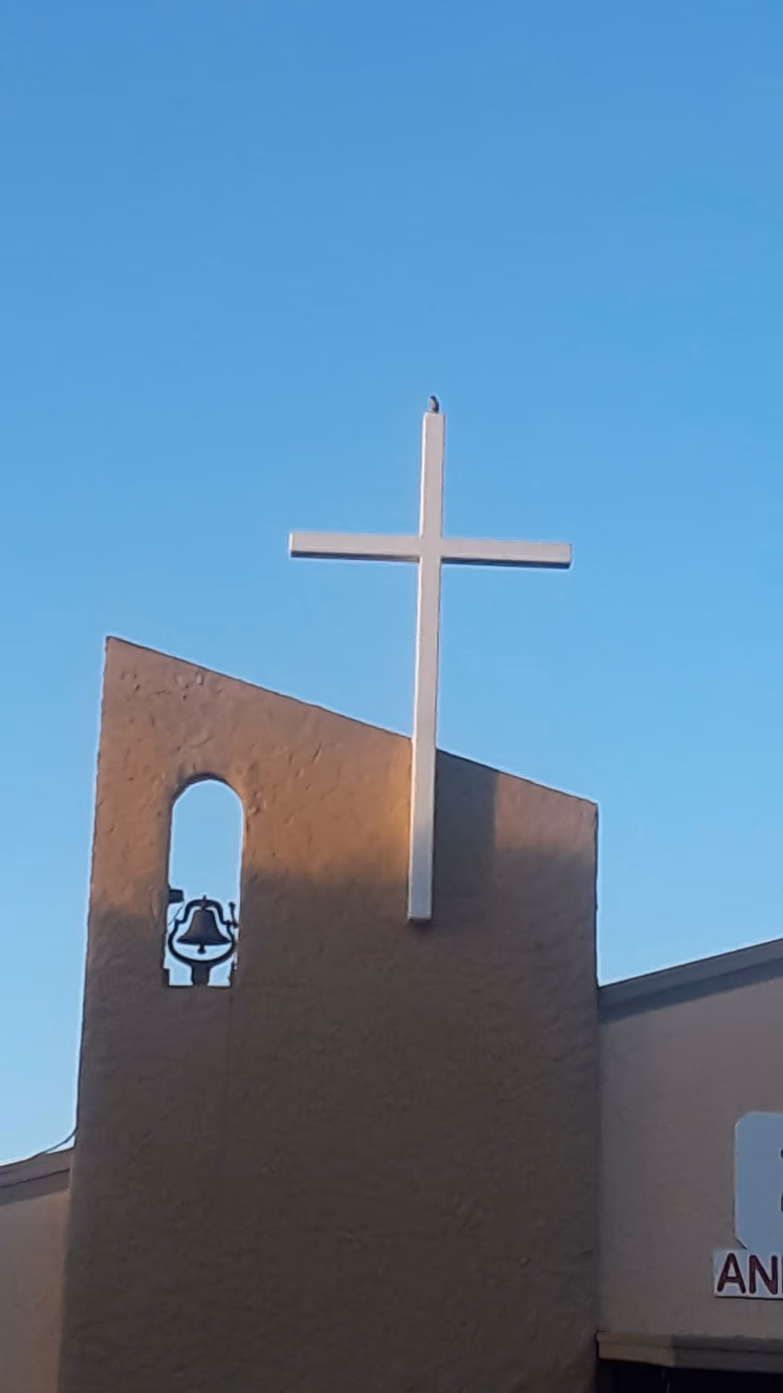 White cross atop a beige building facade with a bell against a blue sky.