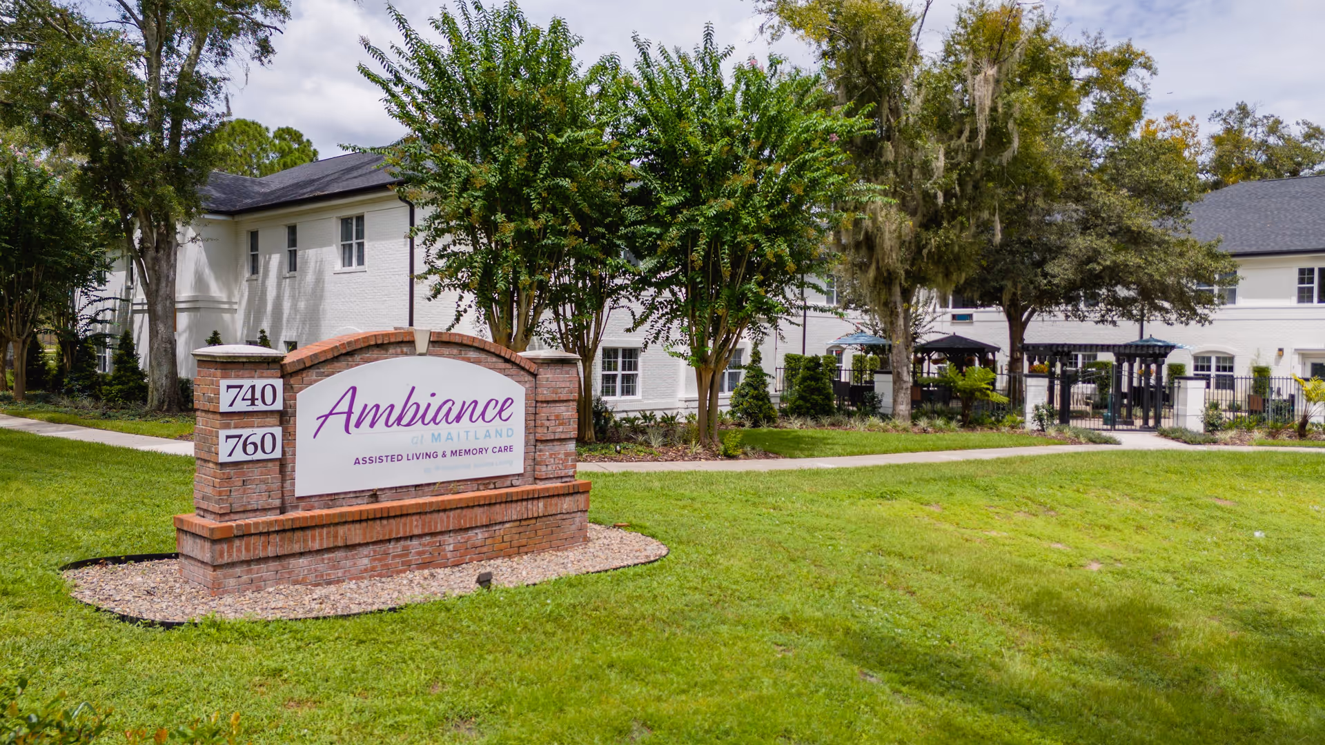 Brick entry sign reading "Ambiance at Maitland" in front of a white two-story assisted living building with lawn, trees, and landscaping.
