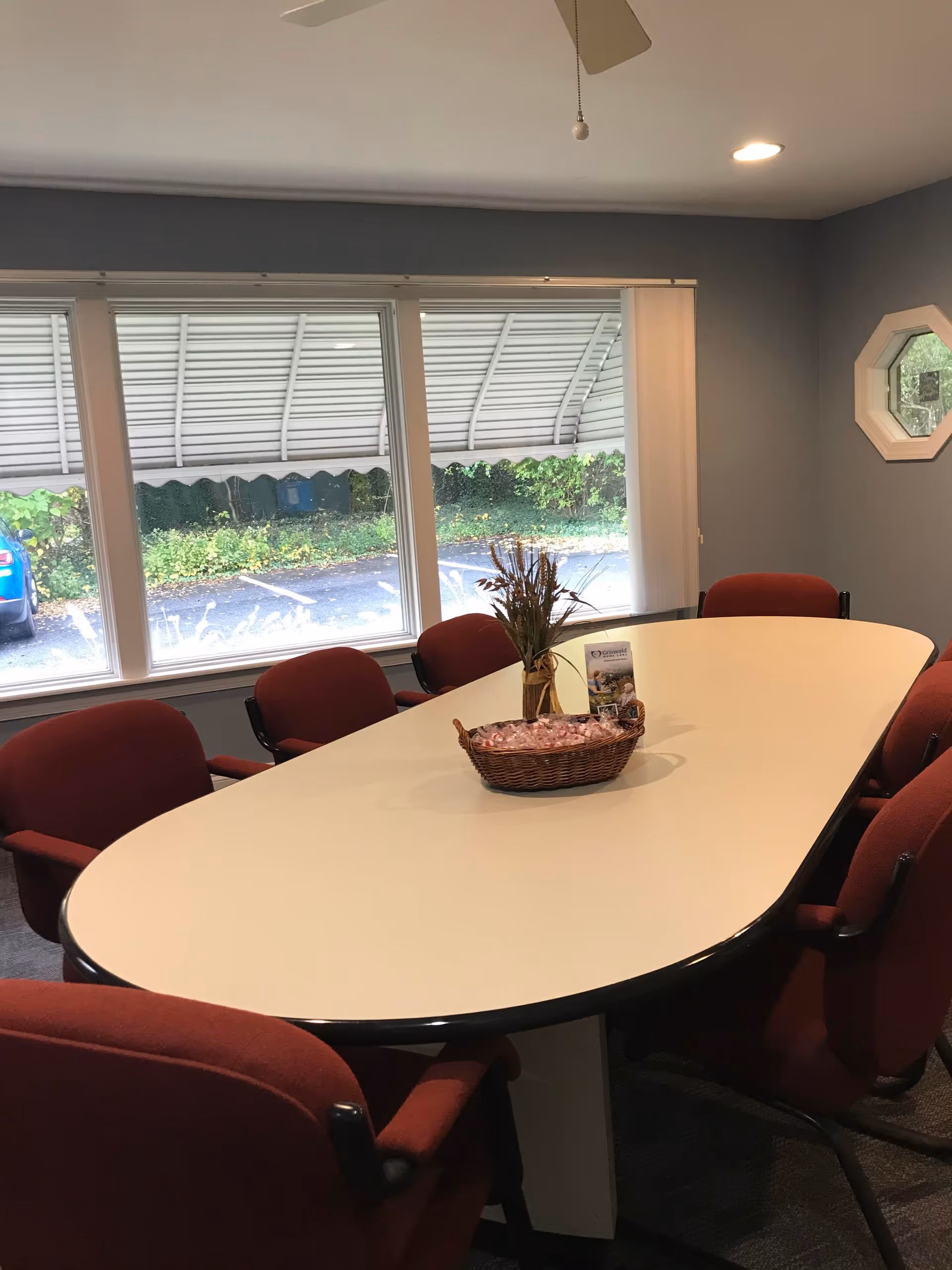 Conference-style room with an oval white table, red chairs, and large windows.