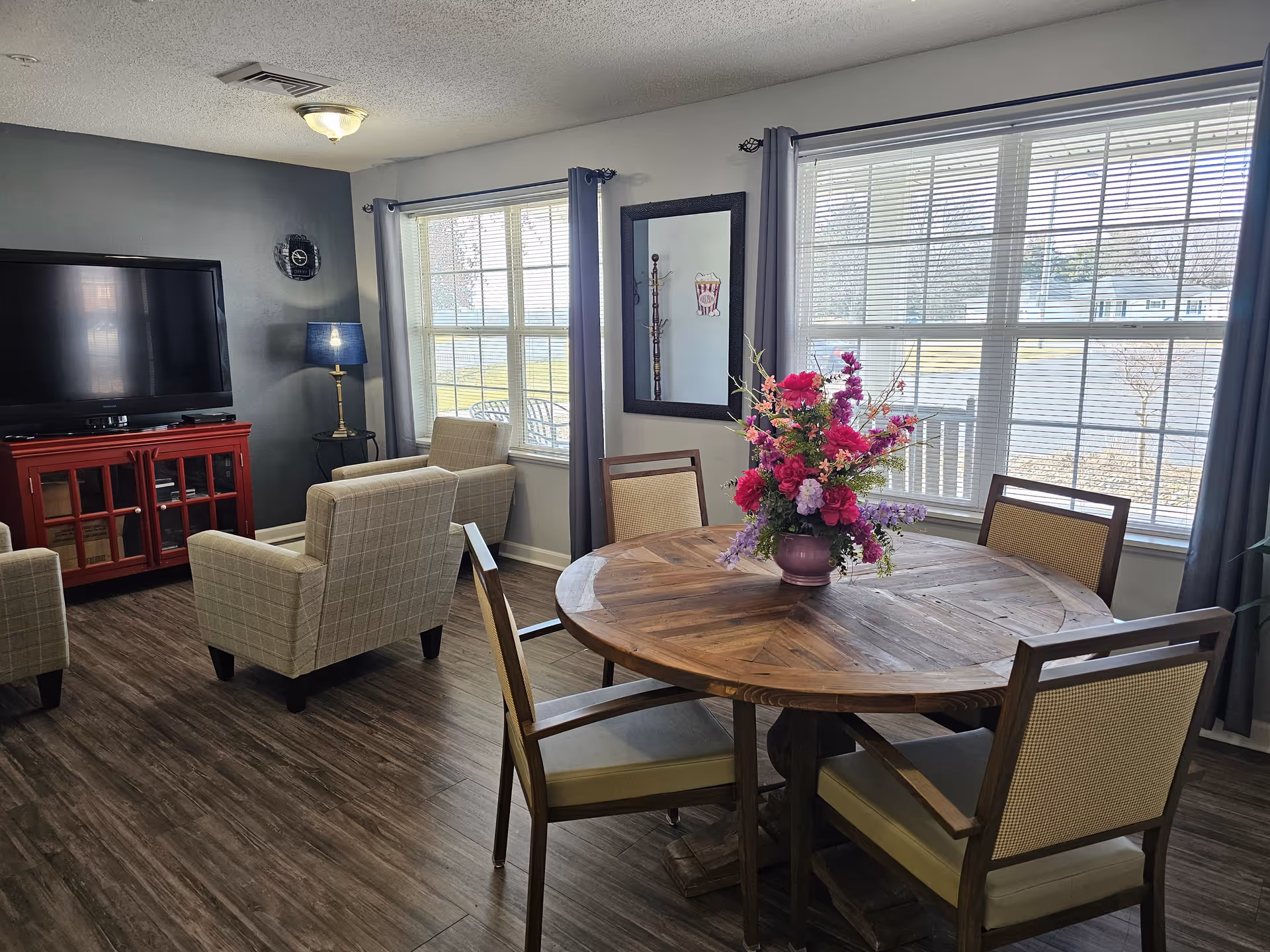 A cozy living area with a round wooden table and four chairs, a vase with colorful flowers on the table, two beige armchairs facing a large flat-screen TV on a red cabinet, a floor lamp with a blue shade, and large windows with gray curtains letting in natural light.