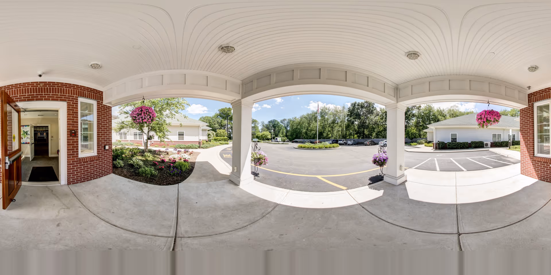 Covered front entrance/portico with hanging flower baskets, brick columns, and a driveway leading to parking and surrounding buildings.
