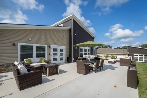 Outdoor patio area at Sugar Maple Commons with wicker furniture including sofas, chairs, and tables. There is a large green umbrella providing shade over a dining table. The building exterior features beige and dark brown siding with white-framed windows and doors. The sky is blue with scattered clouds.