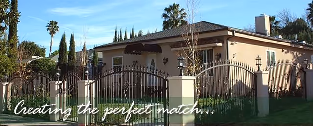 A single-story beige senior living facility exterior behind a gated wrought-iron fence and palm trees.