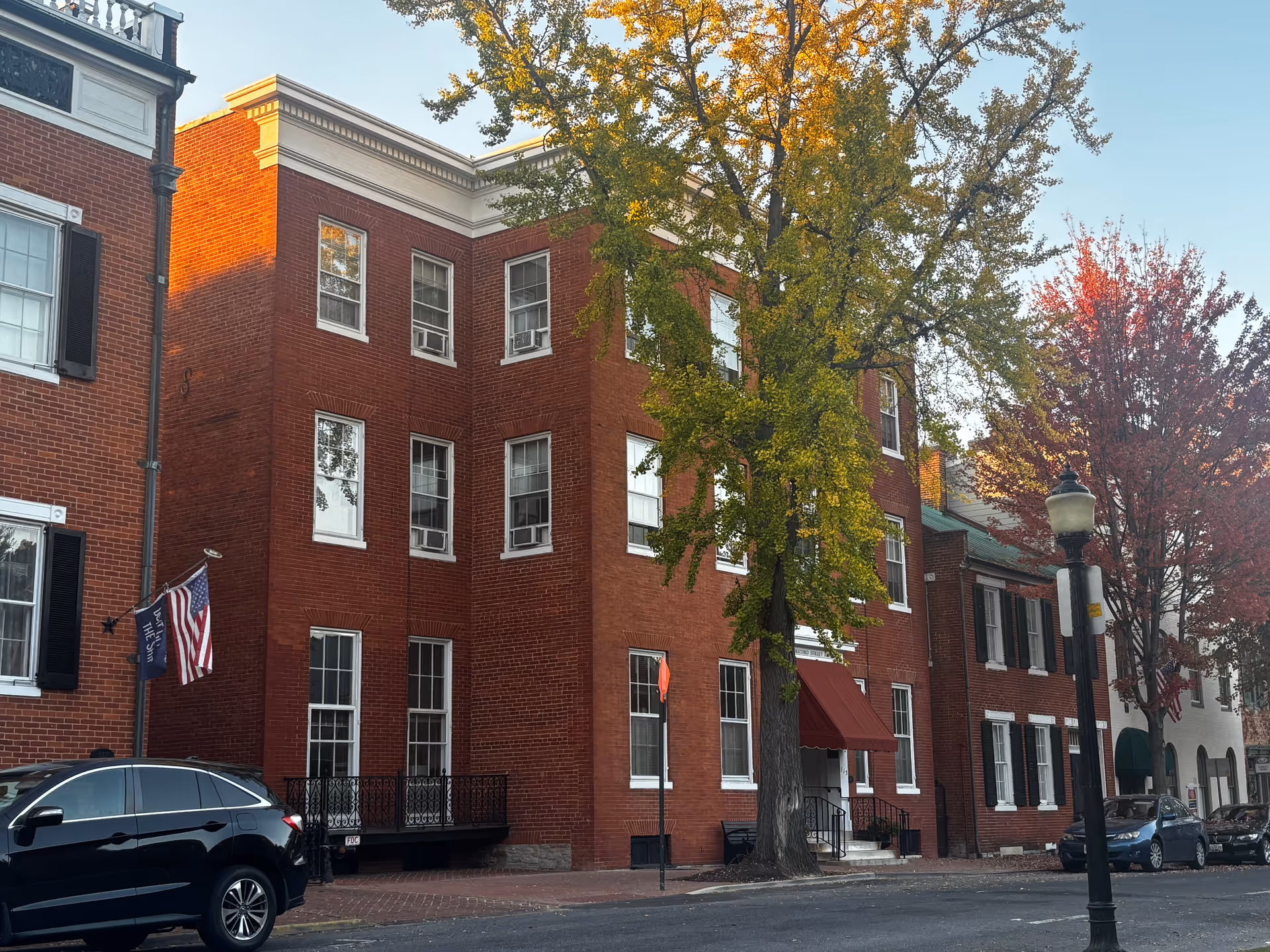 Three-story red brick building with windows and a red awning on a tree-lined street, an American flag, parked cars and a streetlamp in front.
