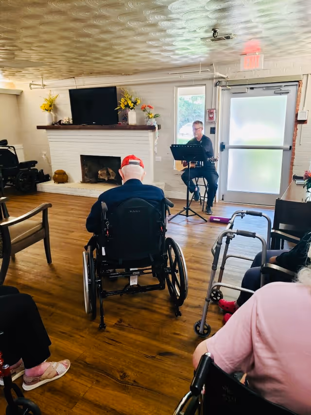 A group of elderly individuals in wheelchairs and with walkers are seated in a common room with wooden floors, facing a man playing guitar and singing near a white brick fireplace decorated with flowers. The room has a TV mounted above the fireplace and a door with frosted glass letting in natural light.