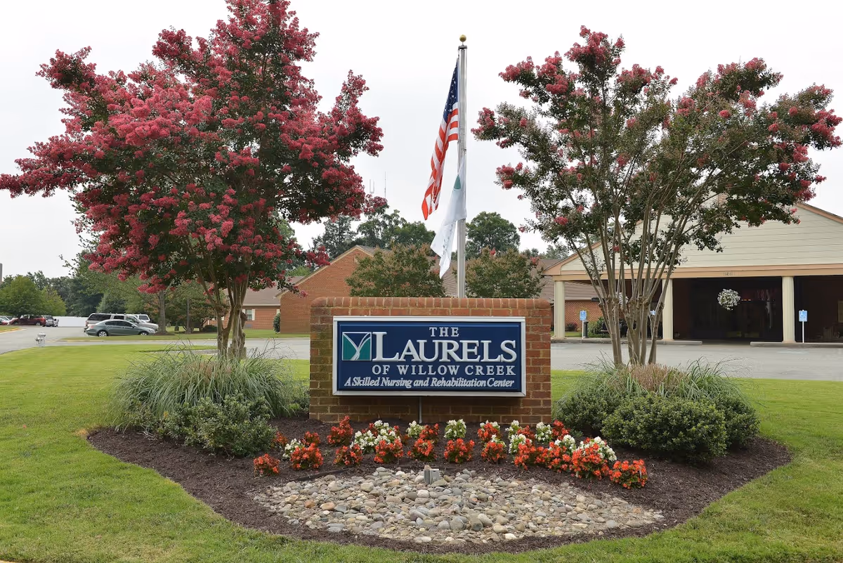Brick sign reading "The Laurels of Willow Creek" at a landscaped entrance with flowering trees and the facility building in the background.