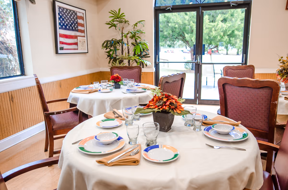 A dining room with round tables covered in white tablecloths, set with plates, bowls, glasses, silverware, and beige napkins. Each table has a floral centerpiece with orange flowers. The room has wooden paneling on the walls, large windows, and glass doors leading outside. There is a framed American flag on the wall and a potted plant in the corner.