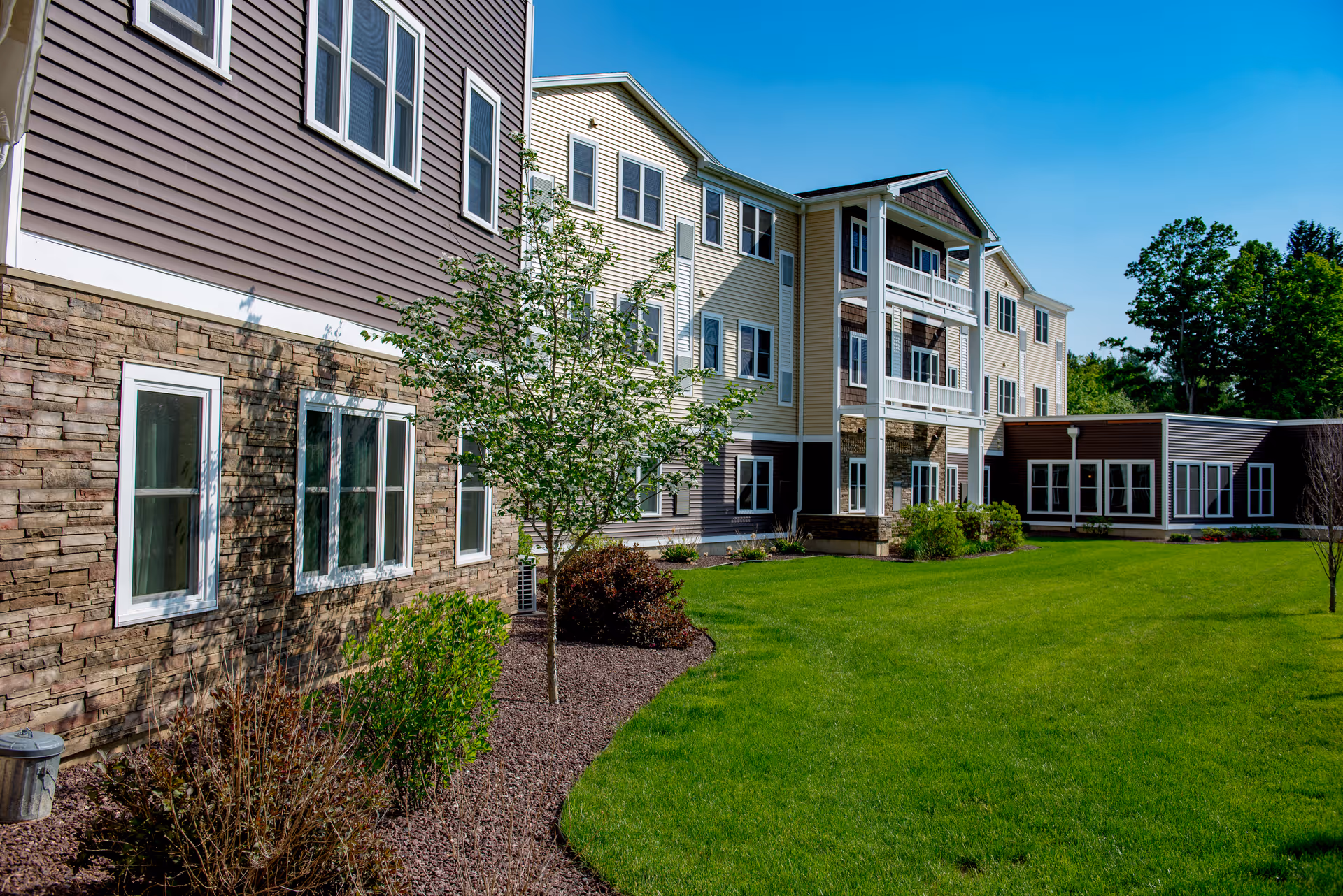 Three-story assisted living building with stone and siding exterior, balconies and a green lawn under a clear blue sky.