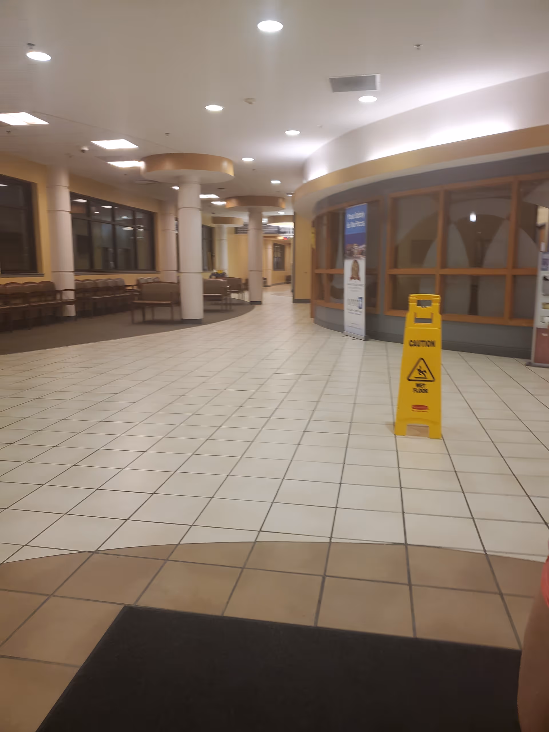Indoor hospital lobby with tiled floors, seating along walls, pillars, and a yellow caution sign.