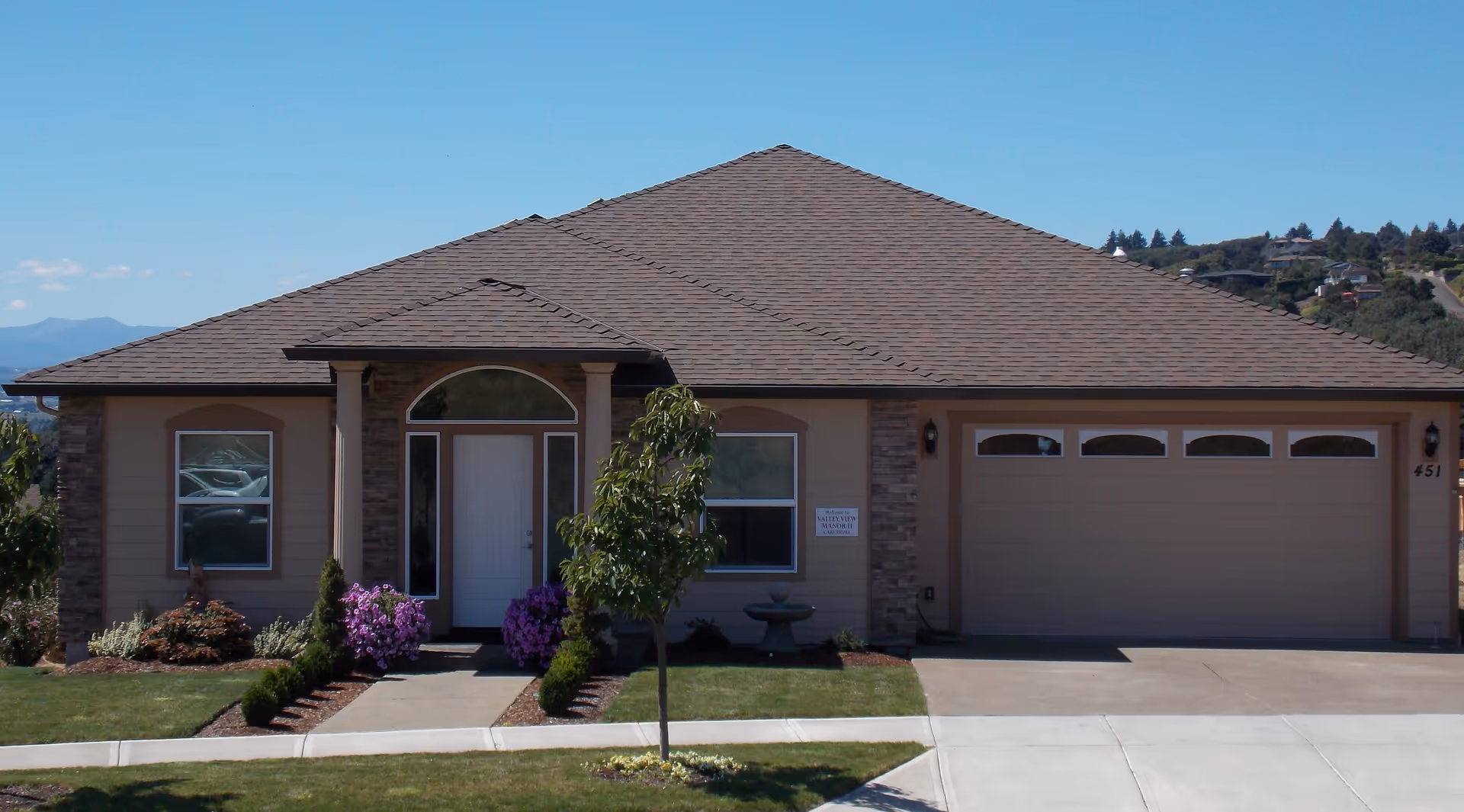 Single-story residential house with a two-car garage, front porch, small lawn and landscaping under a clear blue sky.