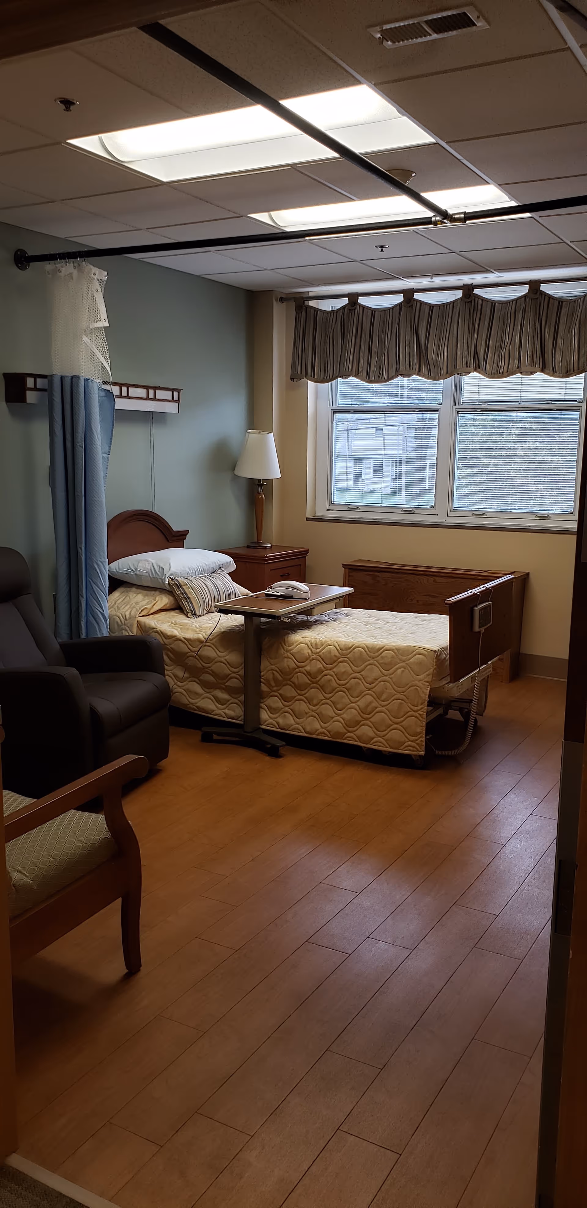 Senior living facility bedroom with bed, chair, nightstand, and large window.