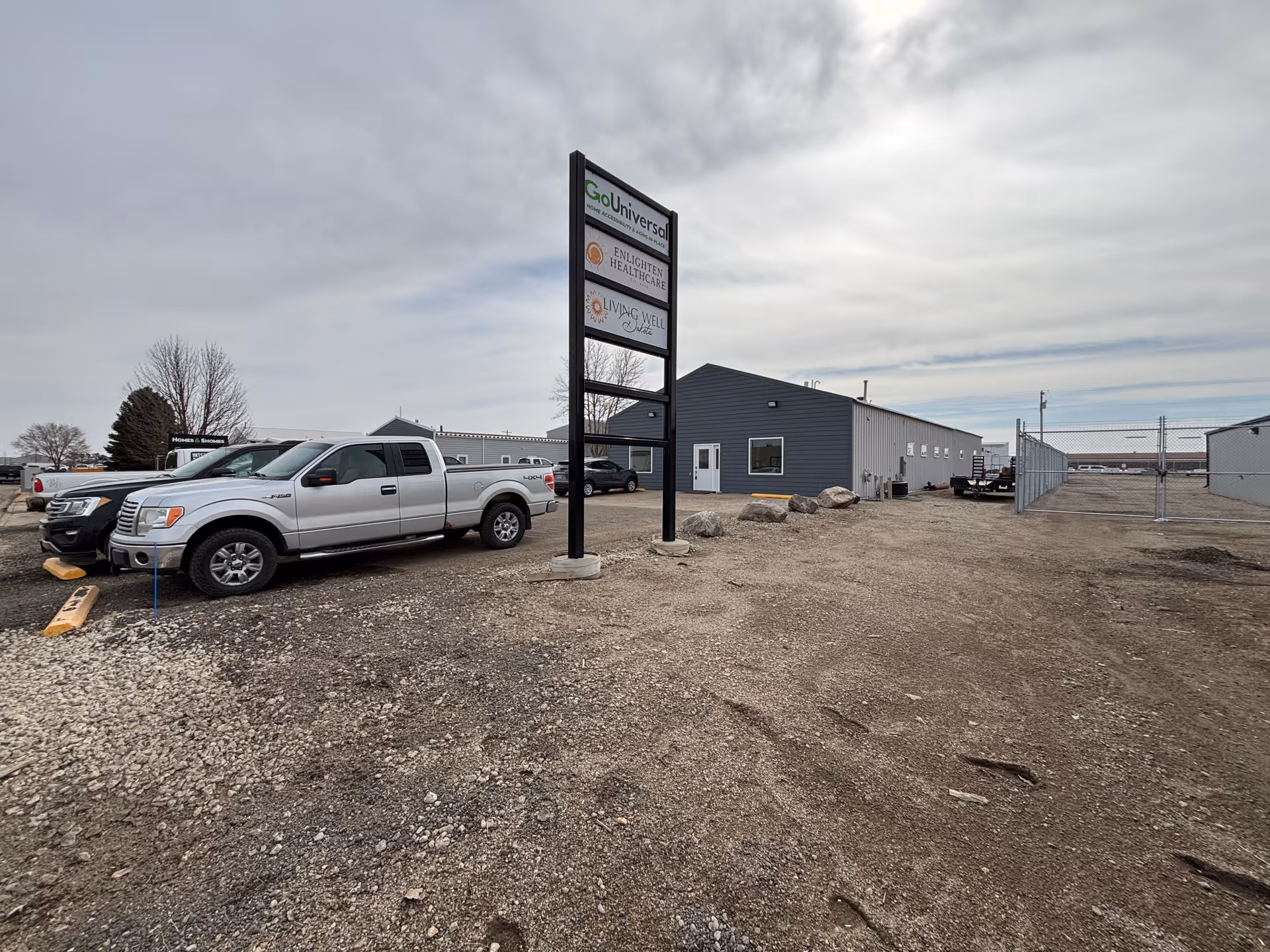 Gravel parking lot with silver trucks, a tall sign for Living Well Dakota, and a building.