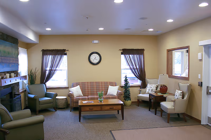 A cozy living room with a plaid sofa, two armchairs, a coffee table, and a side table with a lamp. Two windows with sheer brown curtains flank a wall clock. There is a fireplace on the left side and a small potted plant near the sofa. The walls are painted beige and the floor is carpeted.