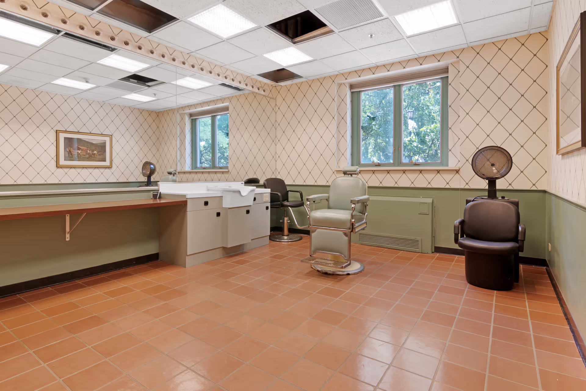 Interior of a salon room with two salon chairs, one green and one black, a hair washing sink, a large mirror on the wall, two windows showing greenery outside, tiled floor, and patterned wallpaper.