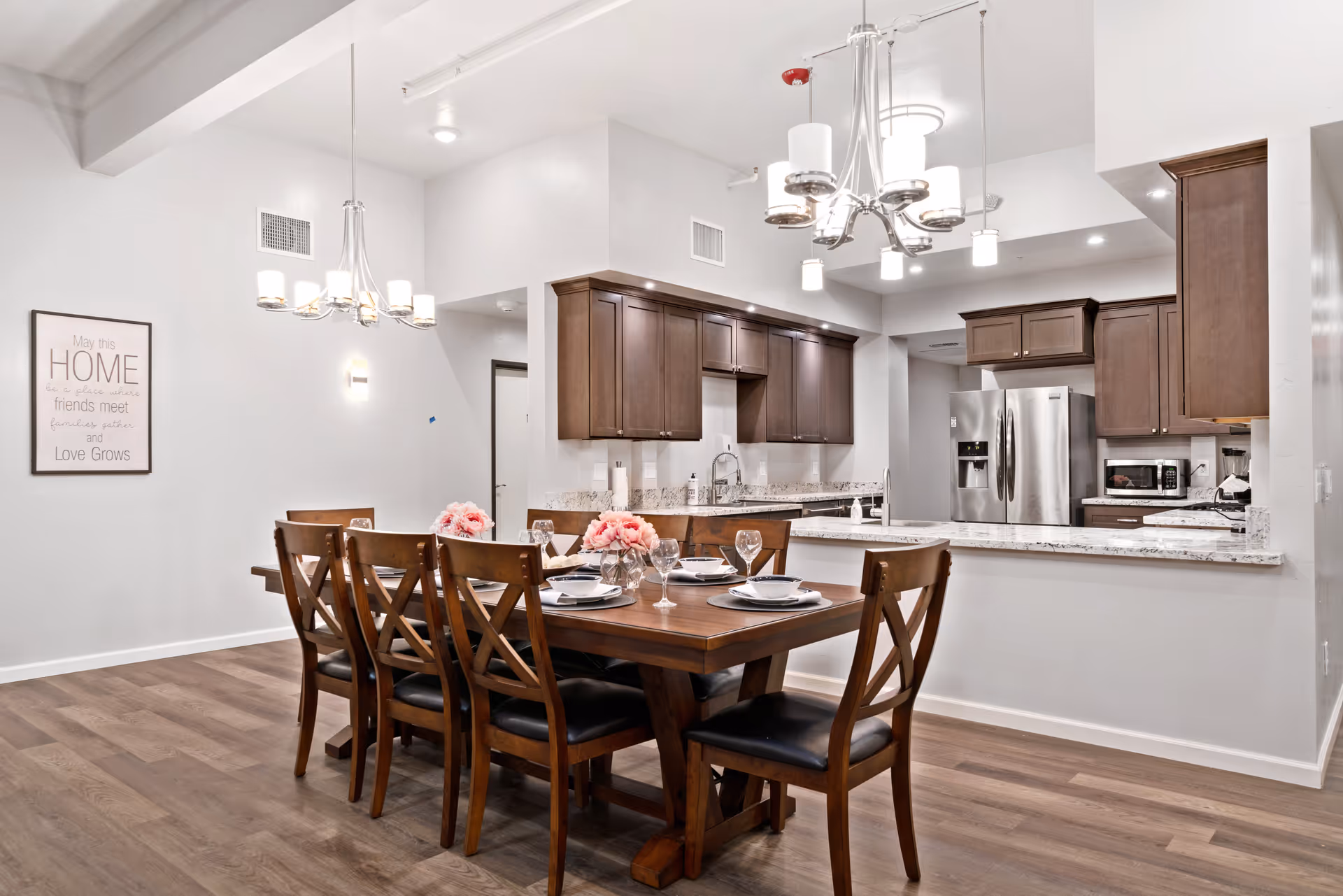 A modern dining area with a wooden dining table set for six, featuring plates, bowls, napkins, and wine glasses. The dining area is adjacent to a kitchen with dark wood cabinets, granite countertops, and stainless steel appliances including a refrigerator, microwave, and blender. Two contemporary chandeliers hang from the ceiling, and a framed wall art with the text 'May this HOME be a place where friends meet, families gather and Love Grows' is visible on the left wall.