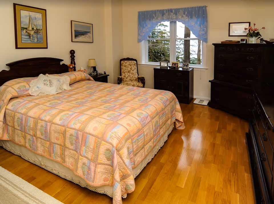 A bedroom with a large bed covered in a pastel patchwork quilt, dark wooden furniture, a window with a blue valance, and hardwood floors.