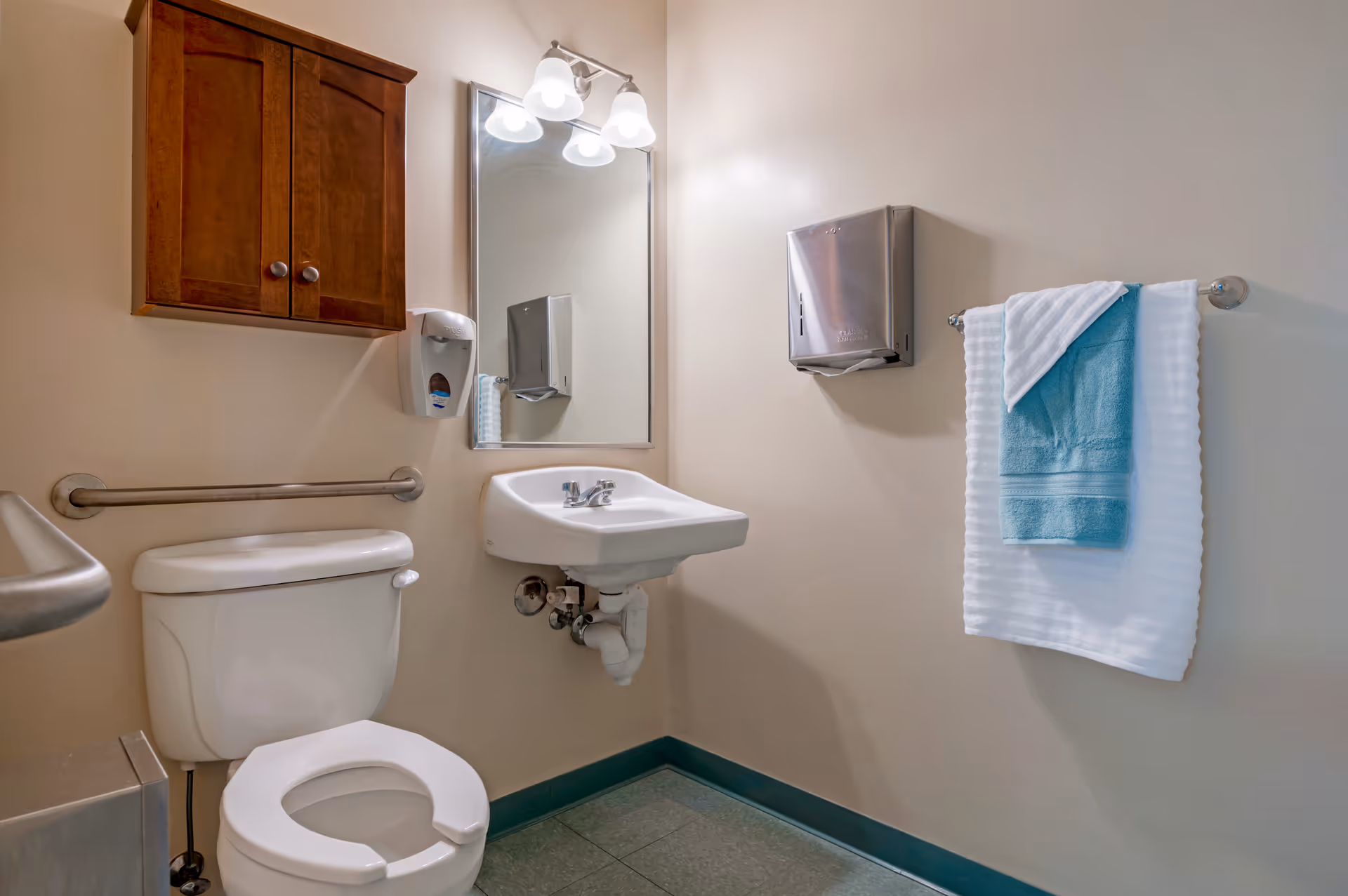 Compact bathroom featuring a toilet, sink, mirror, handrails, and towels.